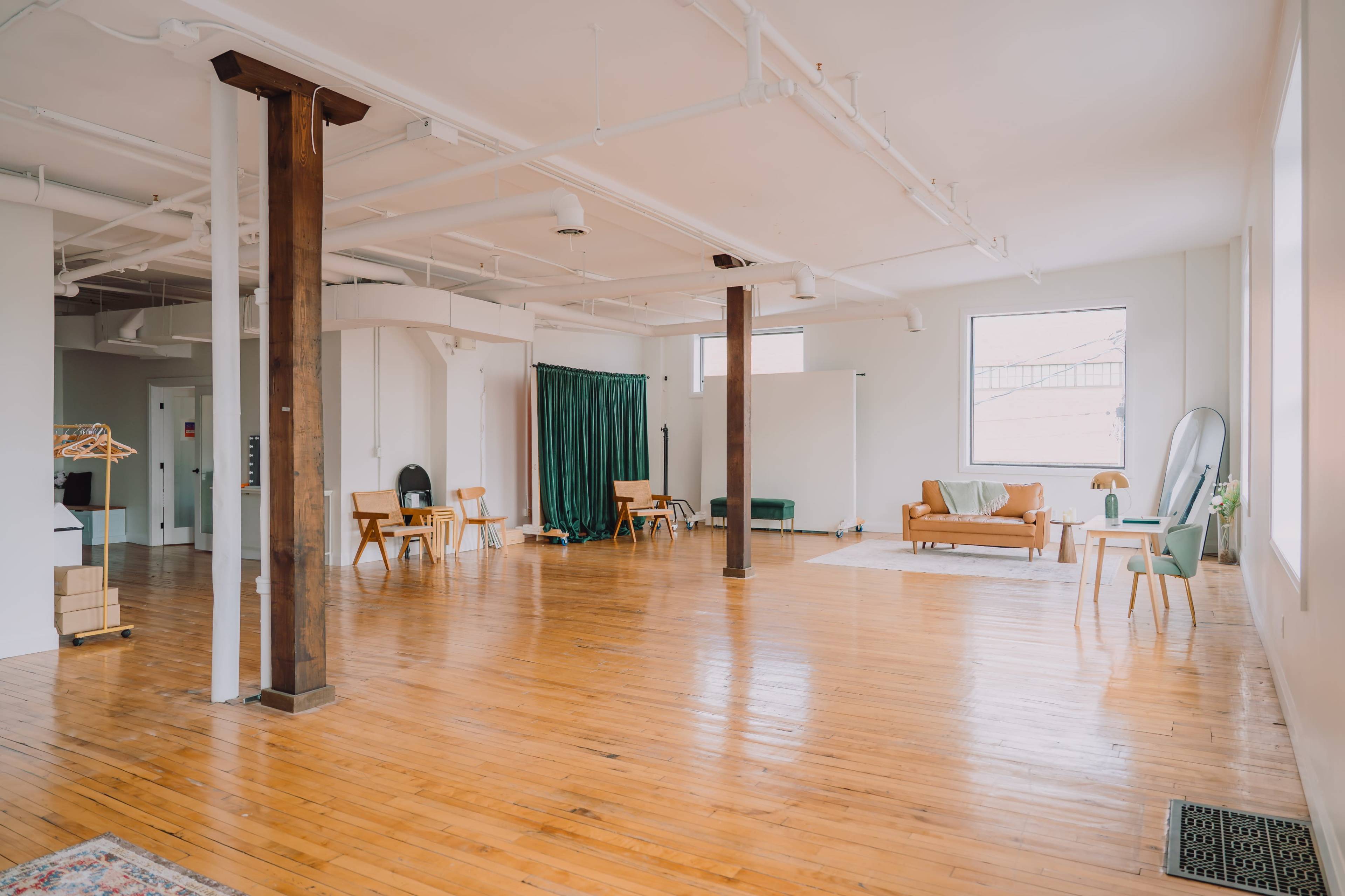 The image shows a spacious, empty loft with wooden floors, large windows, and minimalist furniture including a couch, chairs, and a small table.
