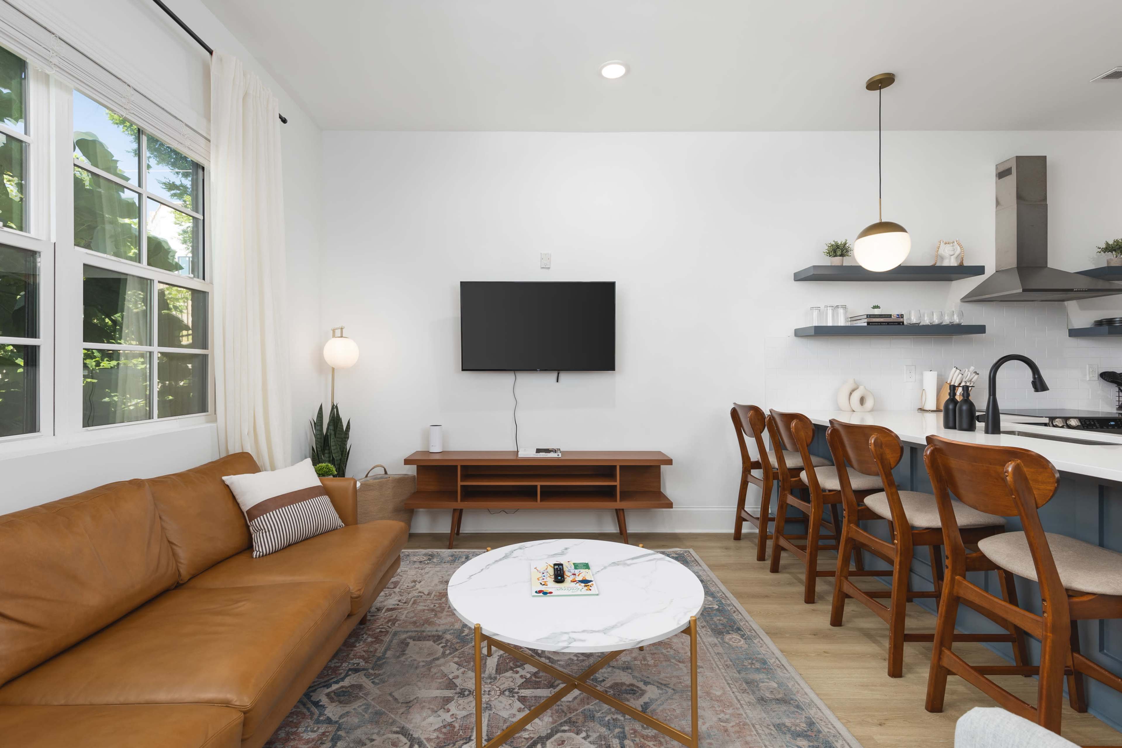 A modern living room features a brown leather sofa, a marble coffee table, and a wall-mounted television, with an adjoining kitchen area that includes wooden bar stools and shelves.