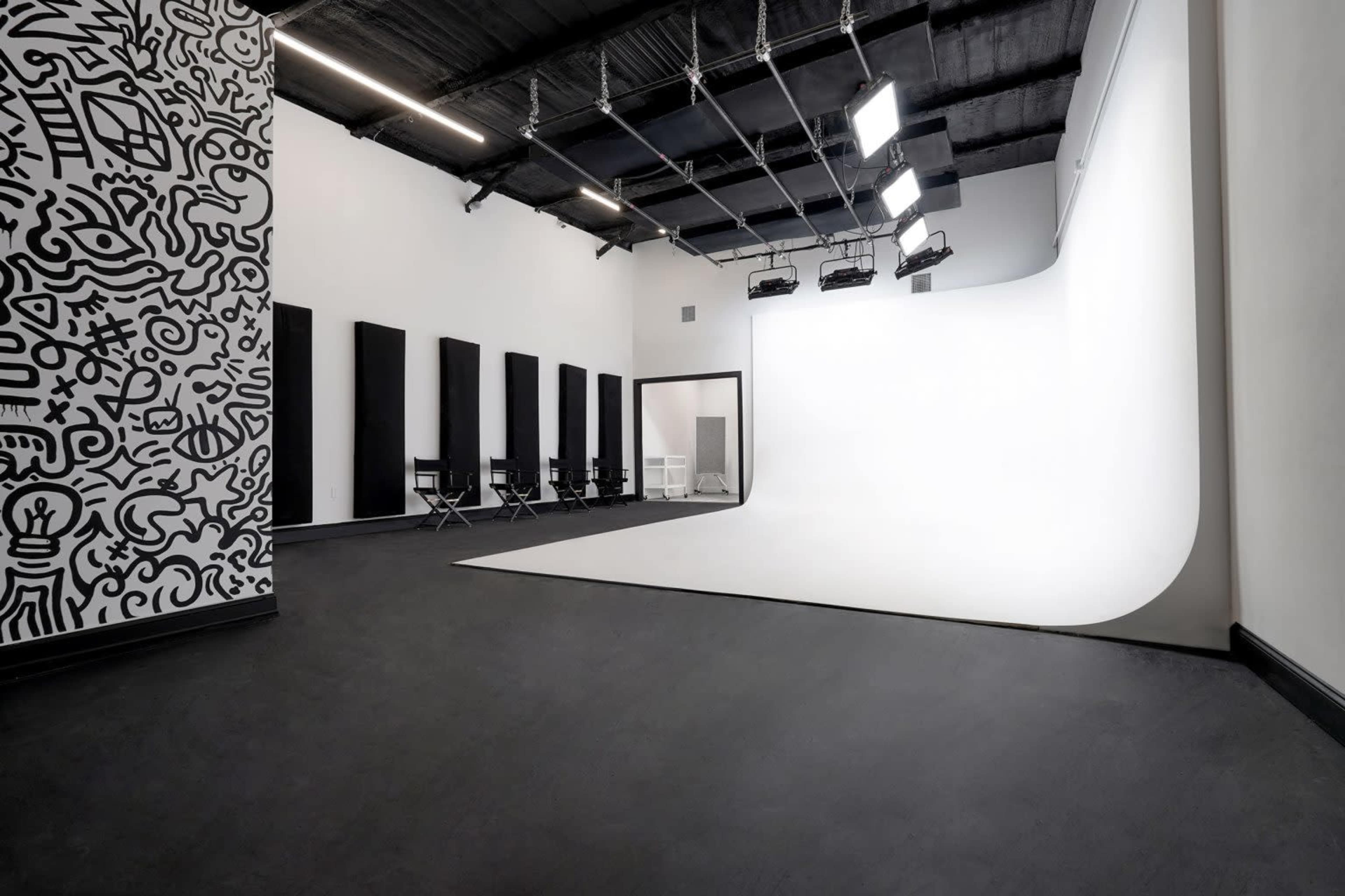 A studio space with a white curved backdrop, black panels on the walls, a patterned mural, and several director's chairs arranged in the corner.