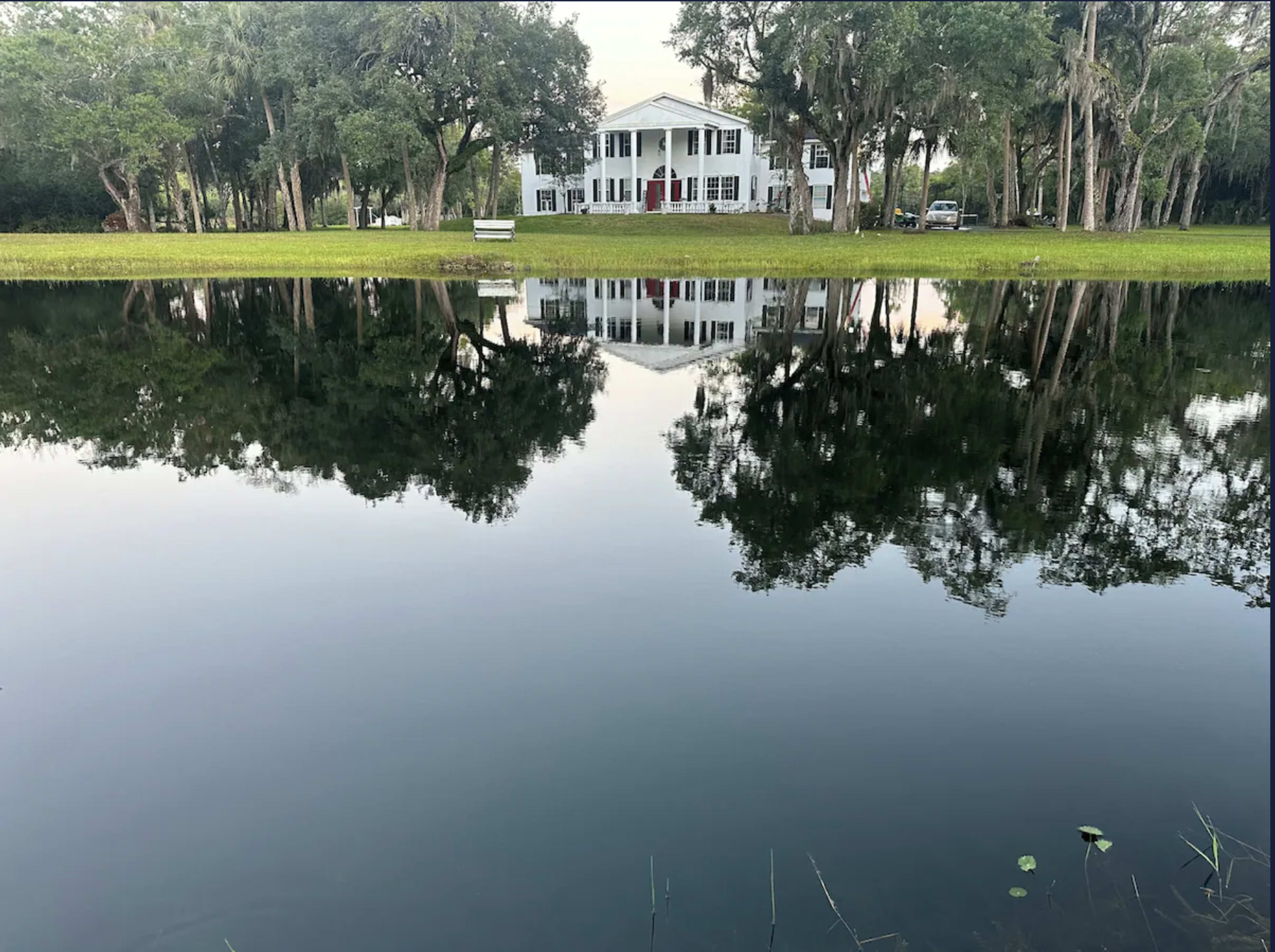 A large white house with a red door stands near a calm pond, reflecting its image and surrounding trees.