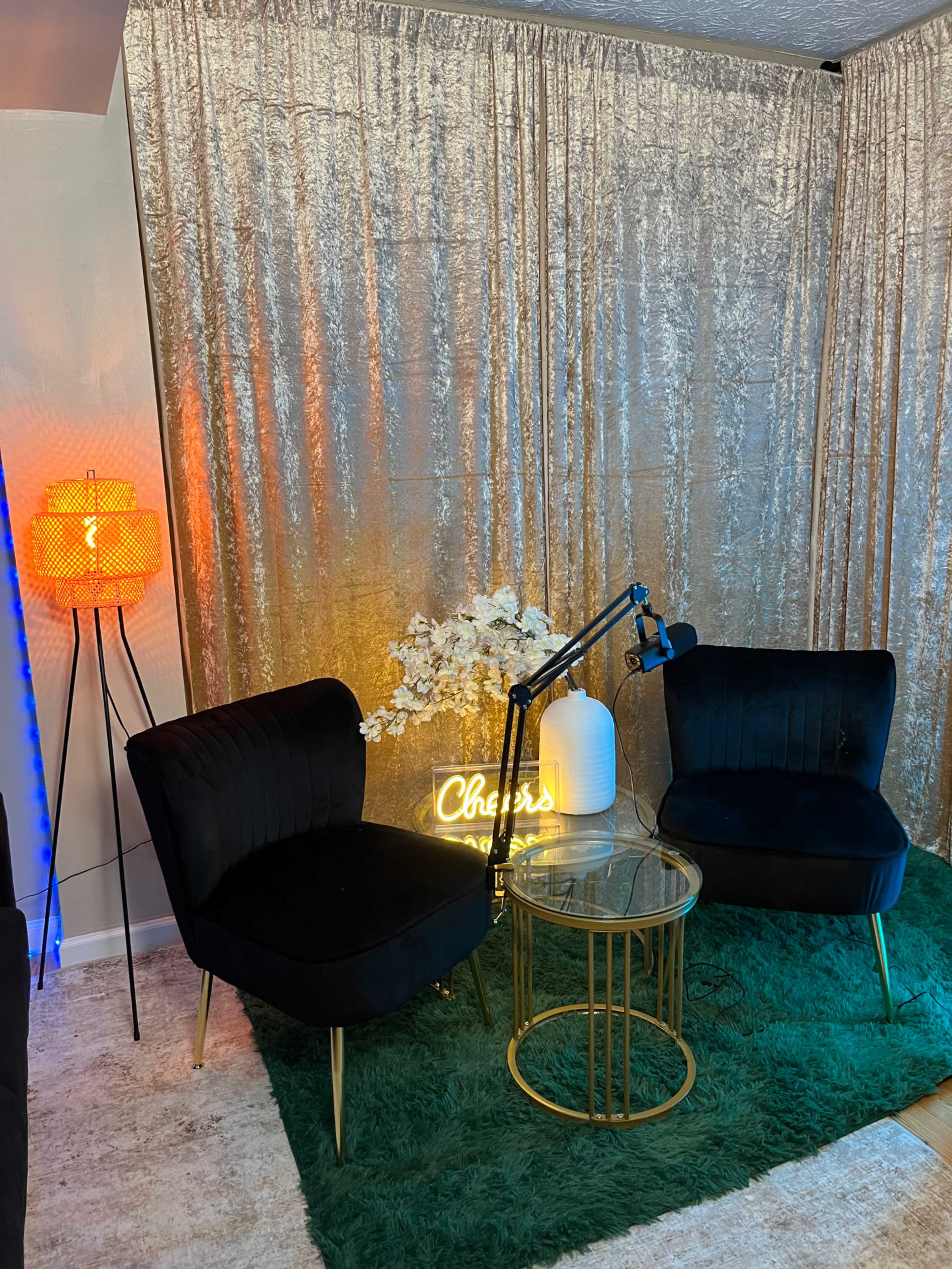 A cozy lounge area features two black velvet chairs, a circular glass table, a decorative lamp, and a gold curtain backdrop with a neon "Cheers" sign.