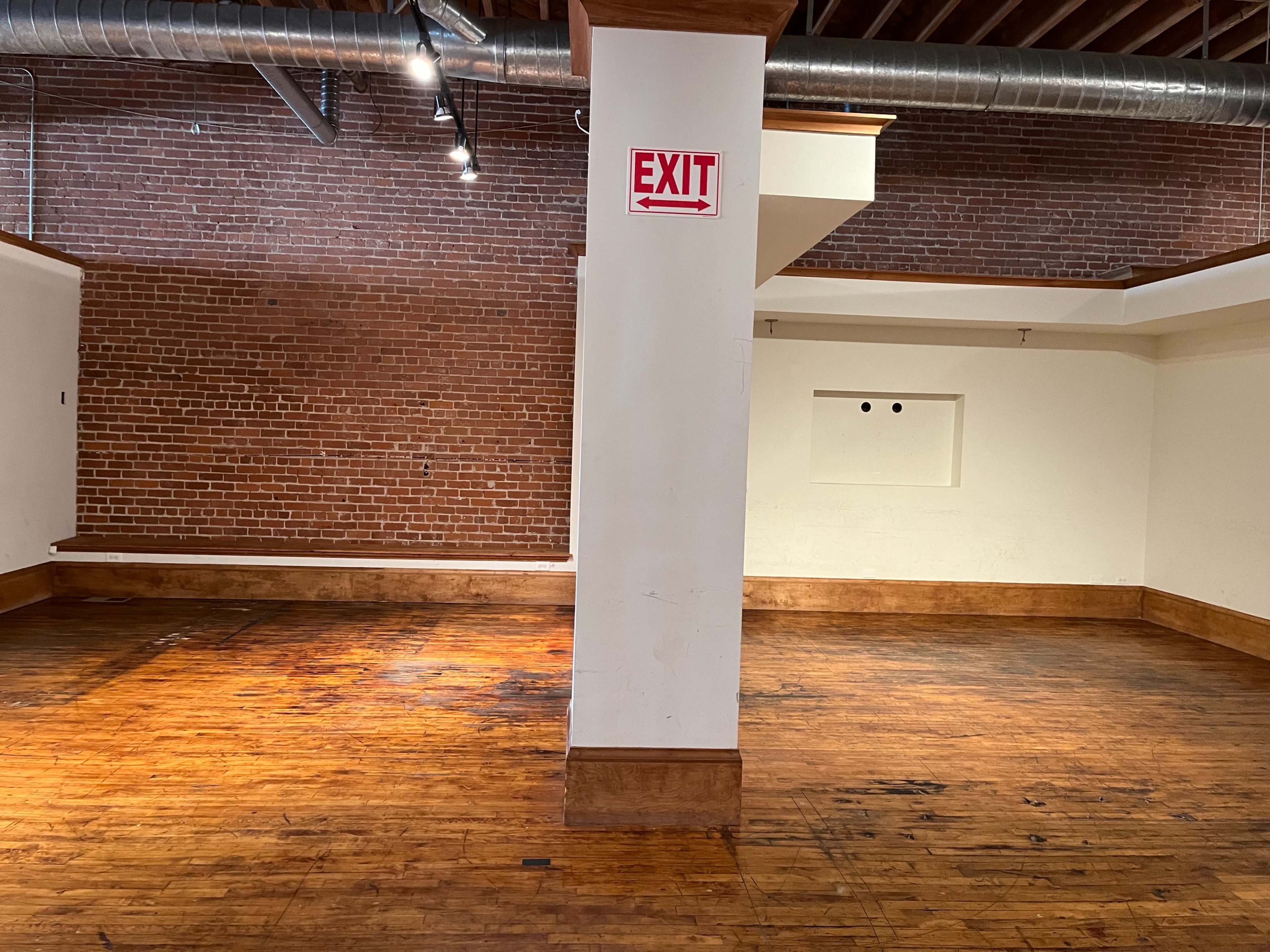 An empty room with wooden floors and exposed brick walls features a central support column and an exit sign hanging on the wall.