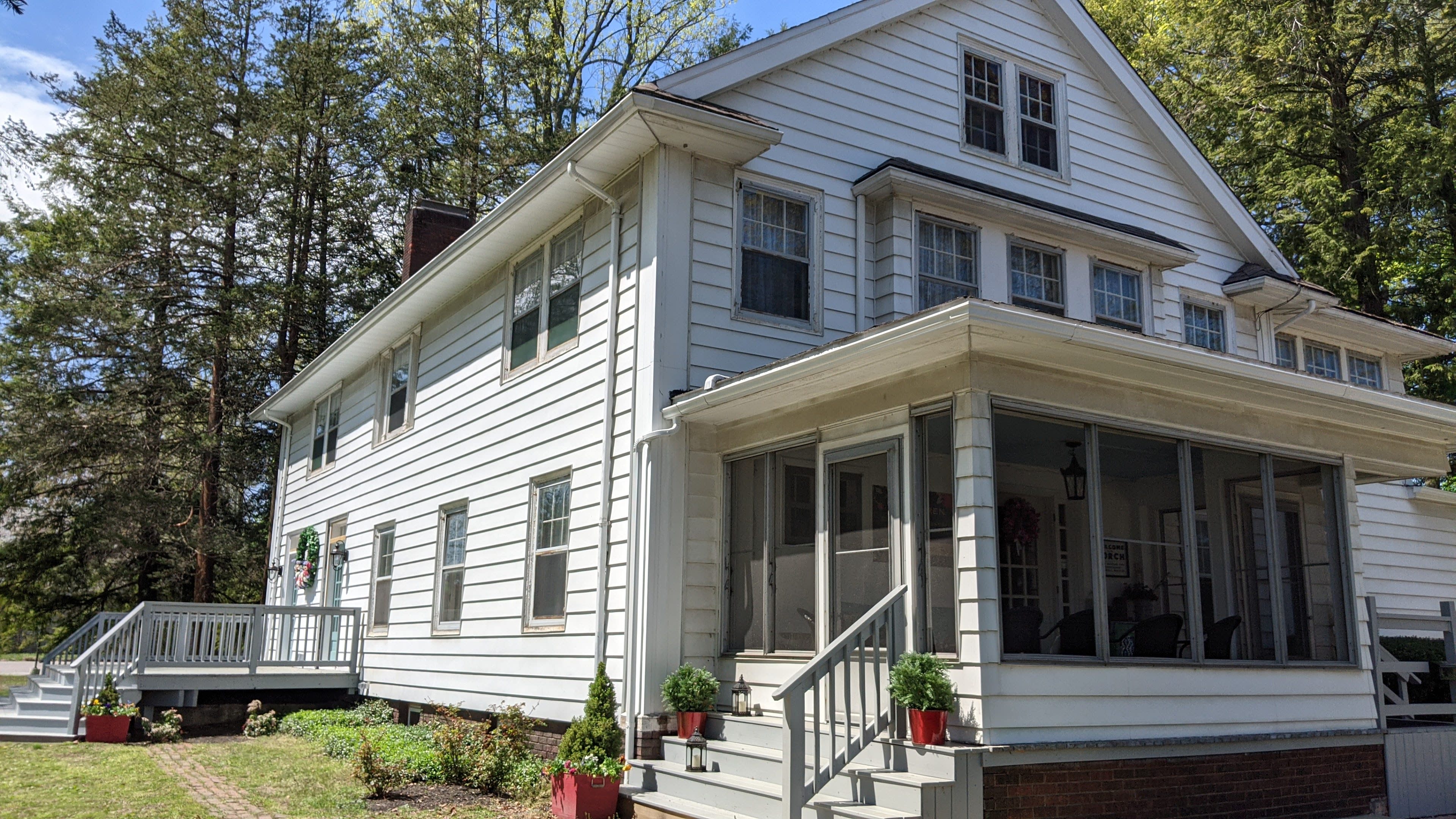 A two-story, white wooden house features a front porch and a side deck, surrounded by greenery.