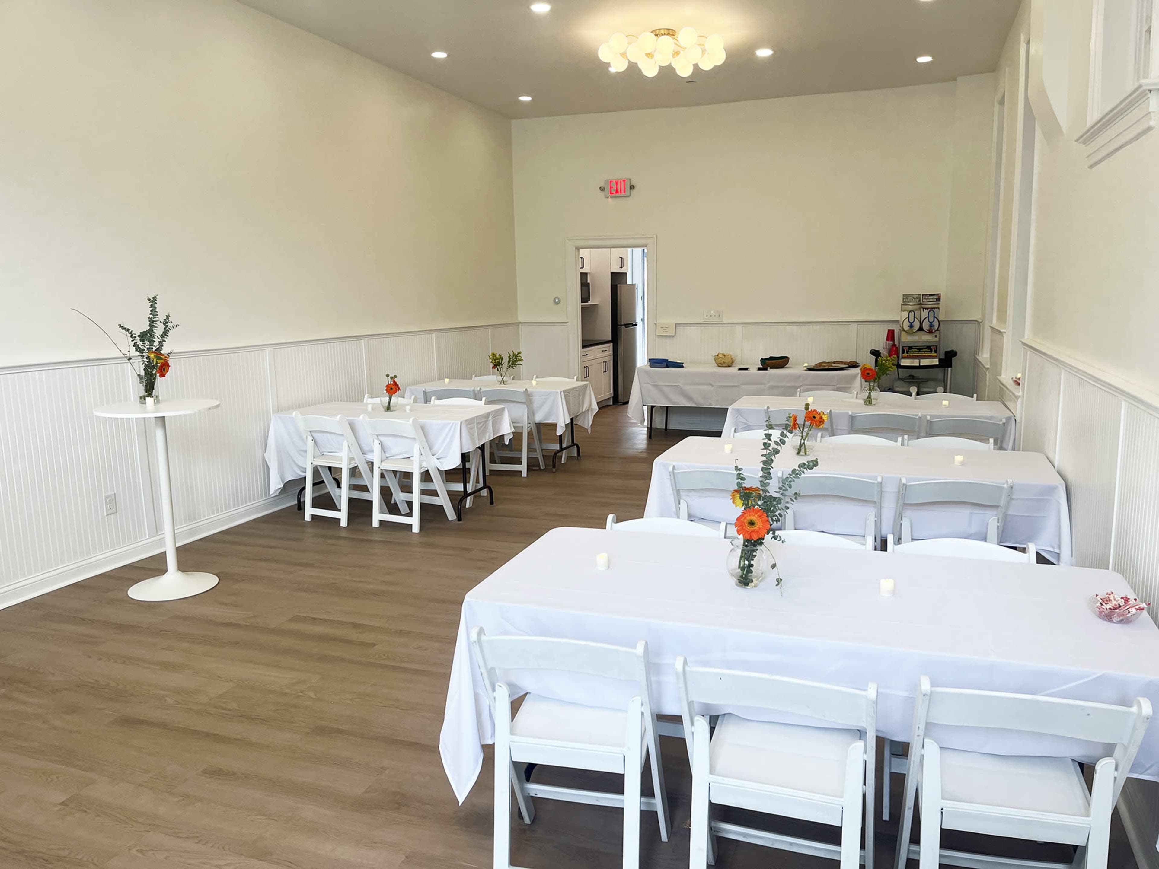 The image shows a clean, well-lit event space with several tables covered in white tablecloths, each adorned with small flower arrangements.