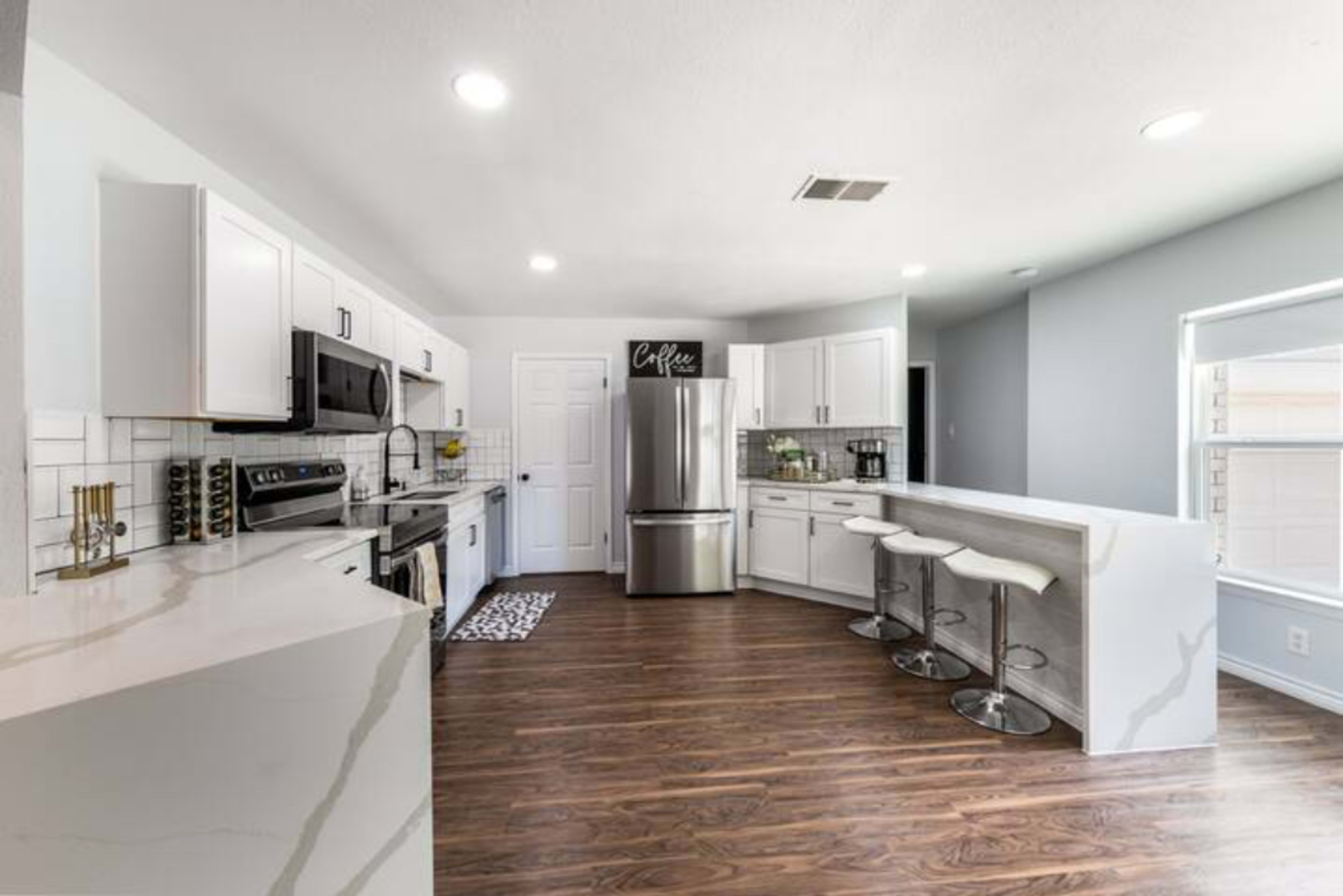 A modern kitchen features stainless steel appliances, white cabinetry, and a bar with three stools.