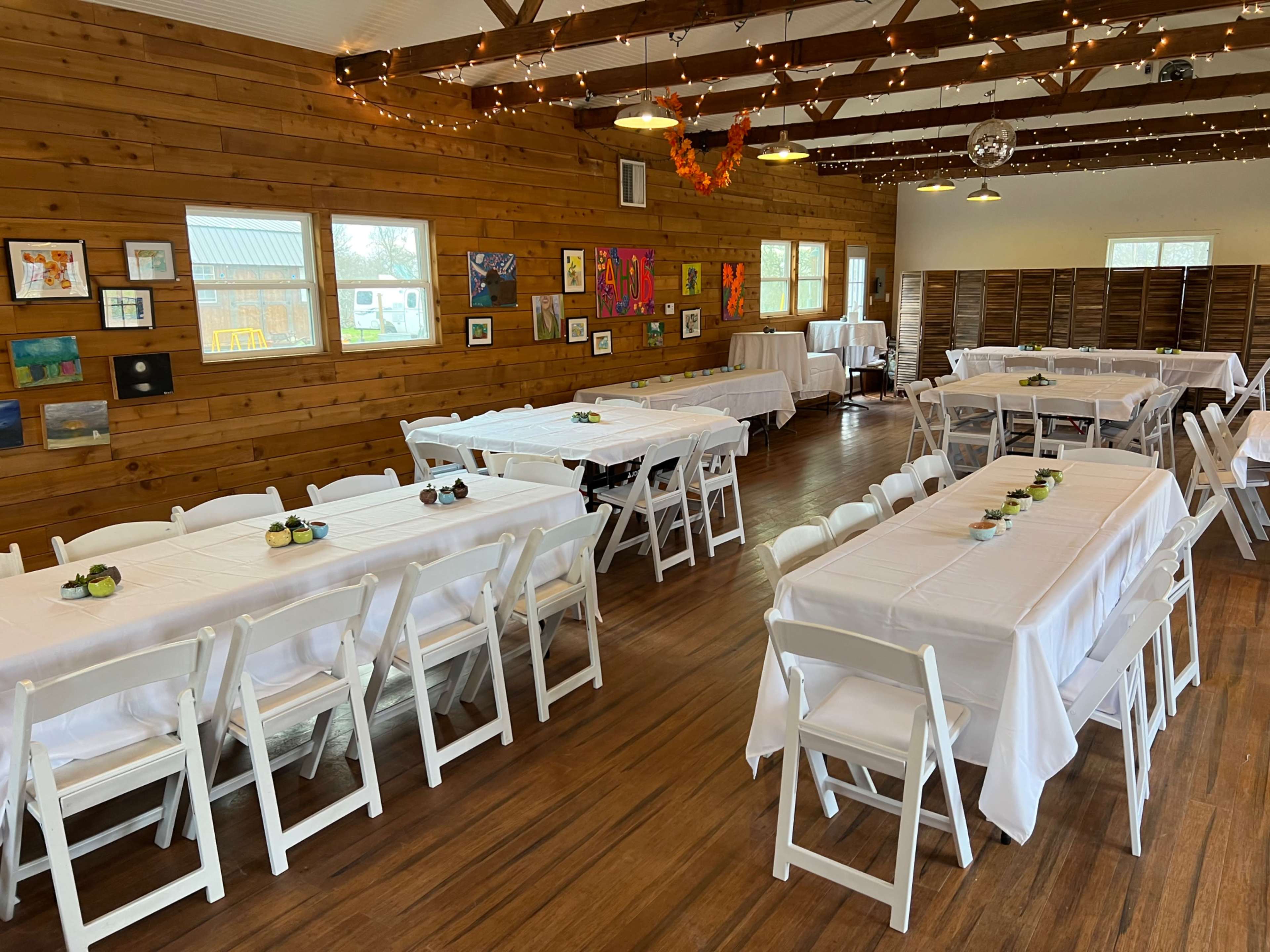 The interior of a banquet hall features neatly arranged tables with white tablecloths and wooden walls adorned with artwork.