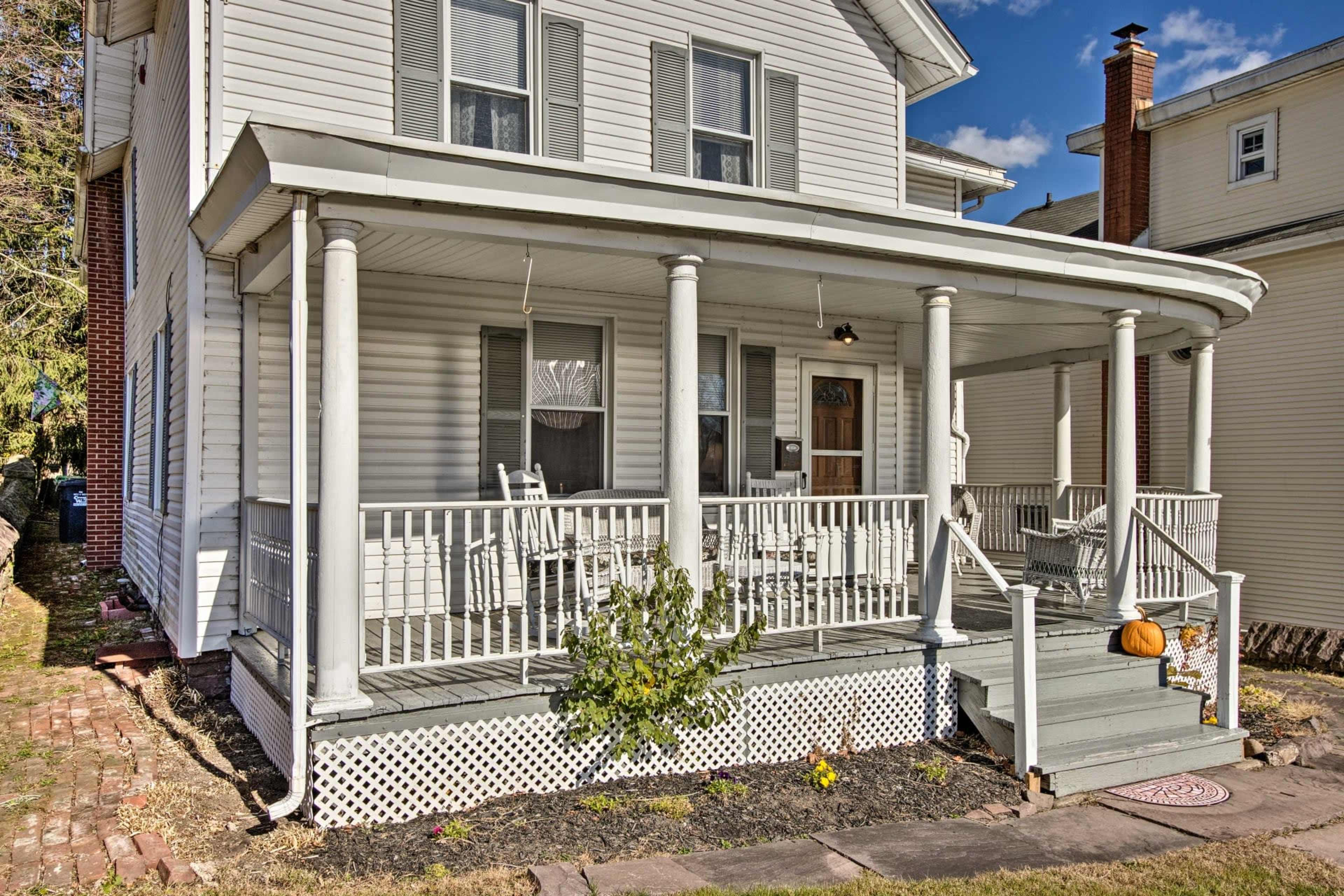 A two-story white house with a covered porch, featuring a railing, two rocking chairs, and a small pumpkin on the steps.