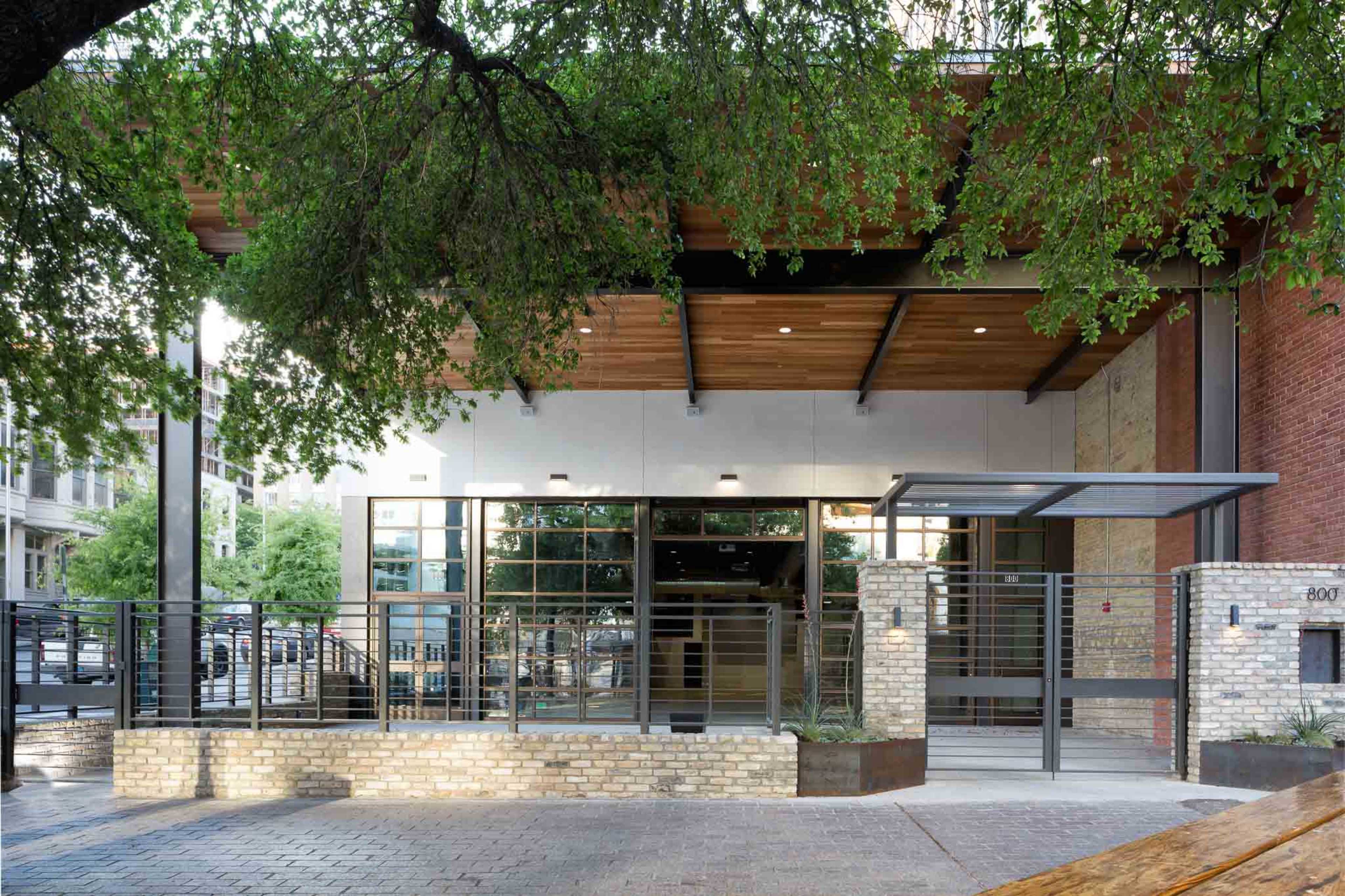 A modern building entrance with large glass doors framed by wooden beams and surrounded by greenery.