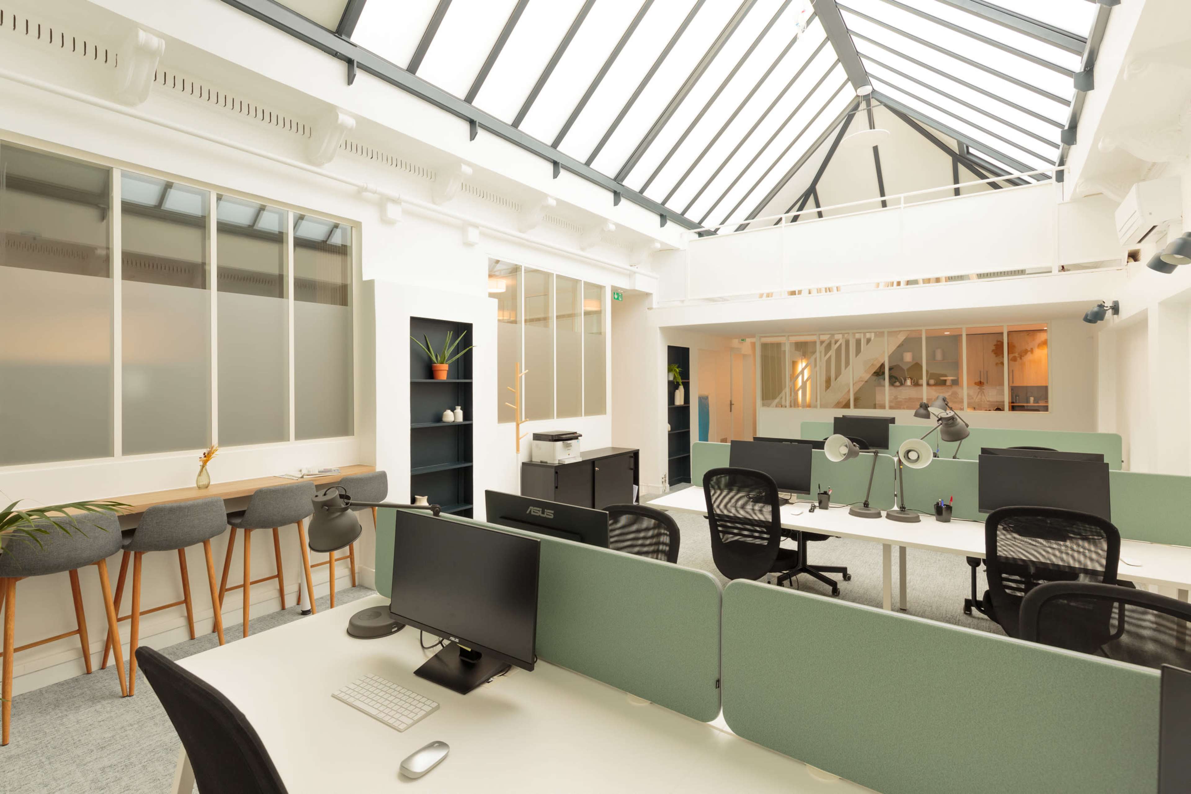 The image shows a modern office space with desks equipped with computers, ergonomic chairs, and a high ceiling featuring glass panels.