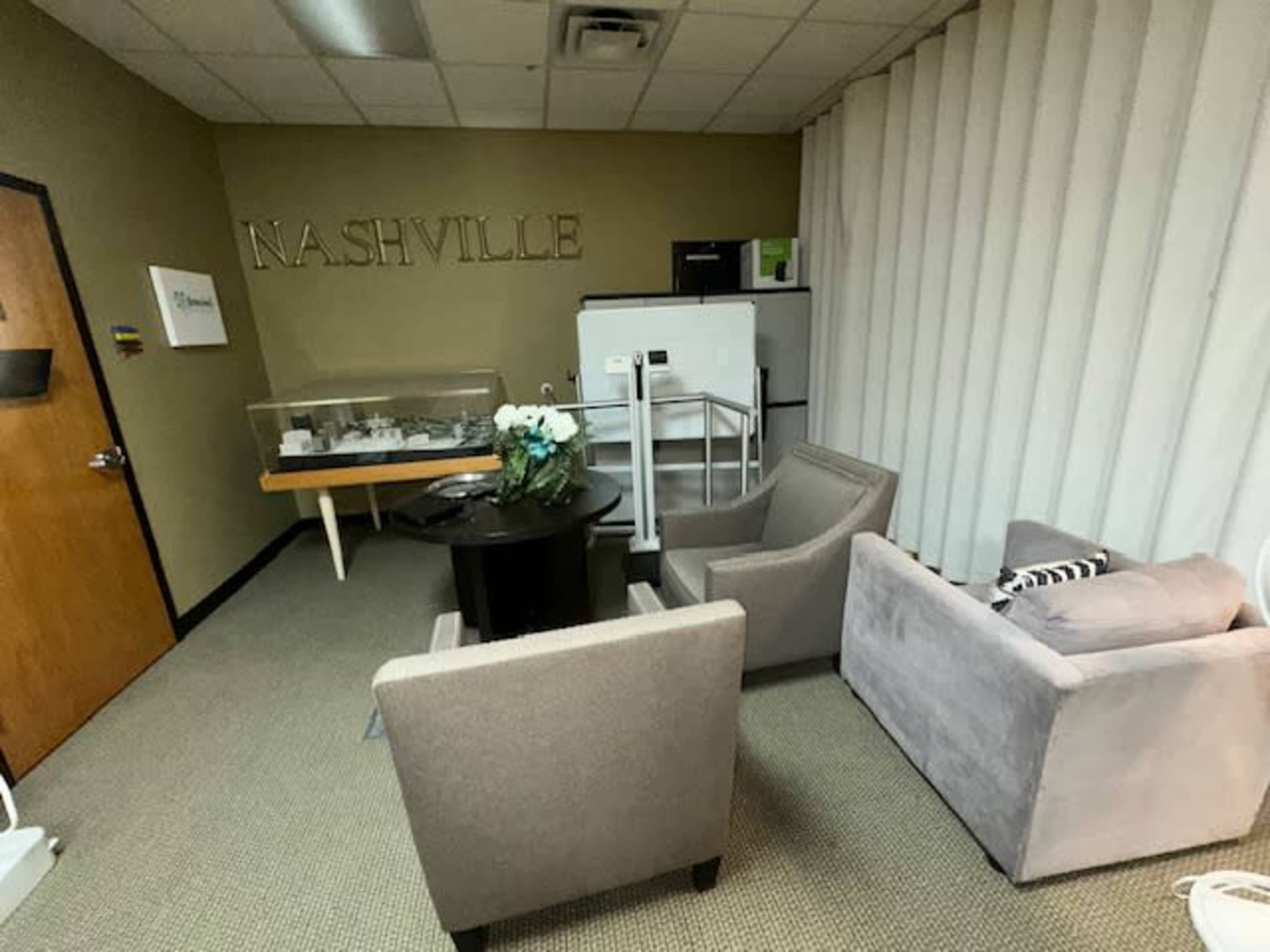 The image shows a small office room featuring a table with a display case, two chairs, and a refrigerator, all against a backdrop of beige walls with the word "NASHVILLE" on one side.