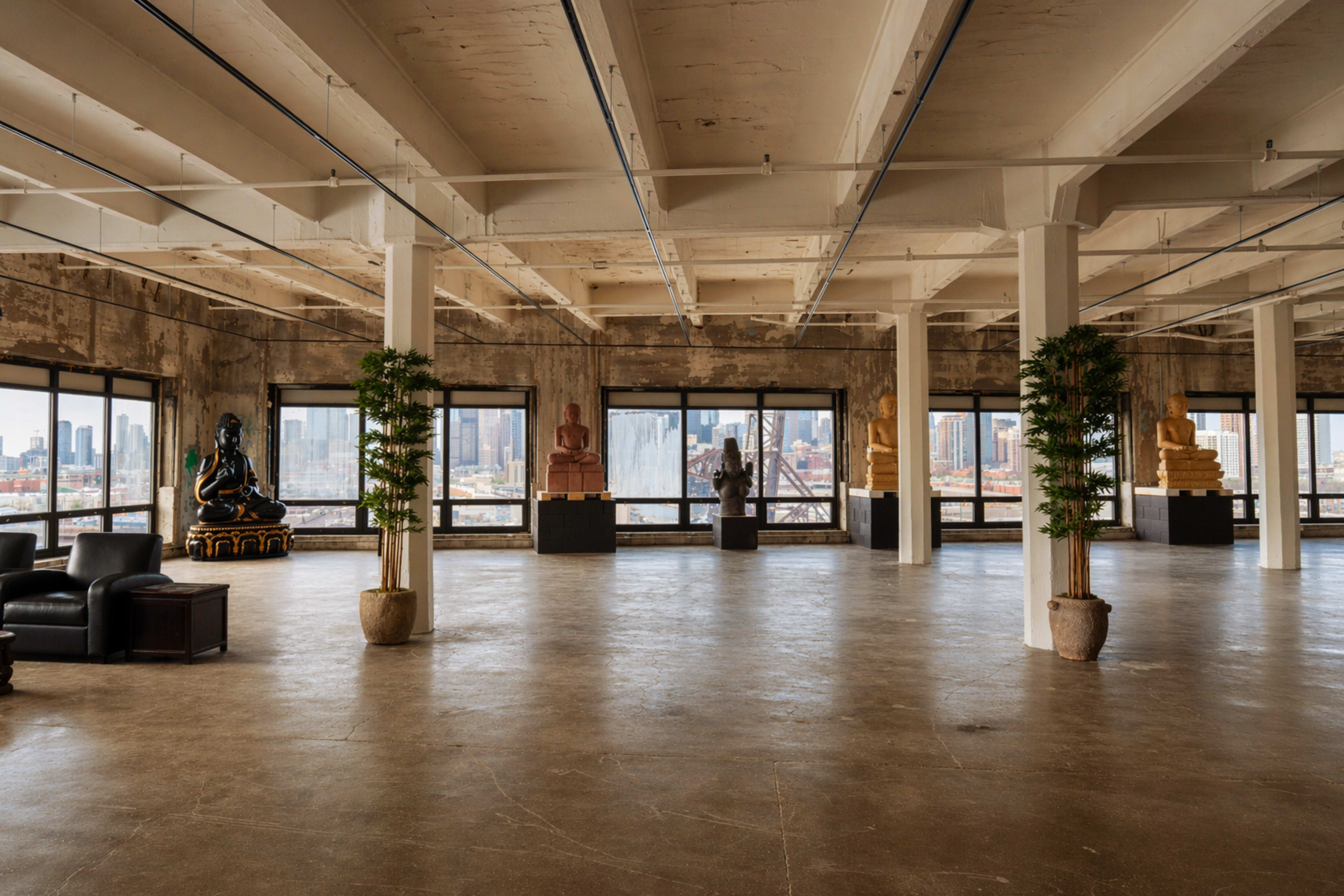 The image shows an expansive, industrial-style loft featuring large windows with city views, adorned with sculptures and potted plants.