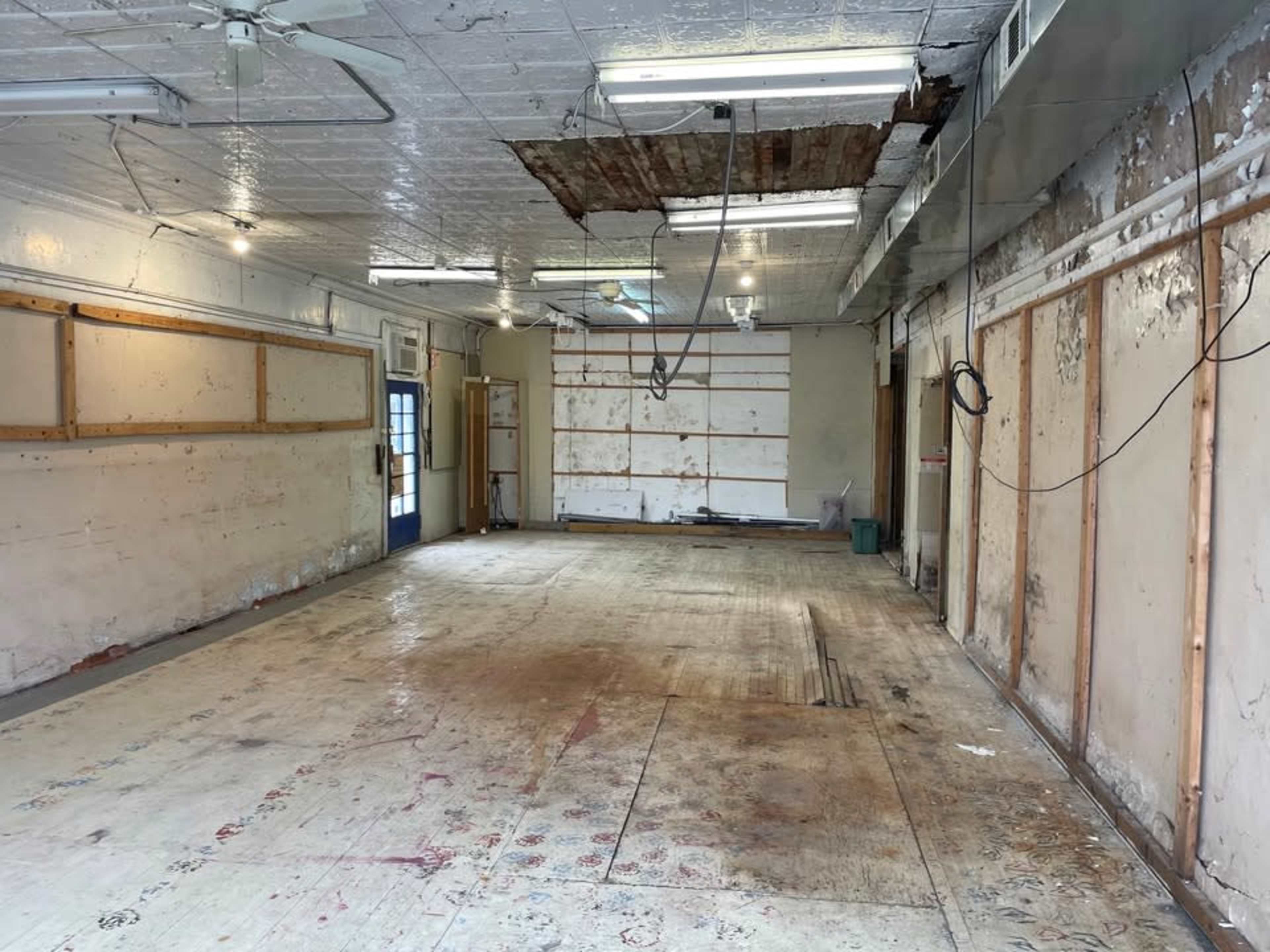 The image shows a large, empty room with a damaged ceiling, exposed walls, and a bare wooden floor.