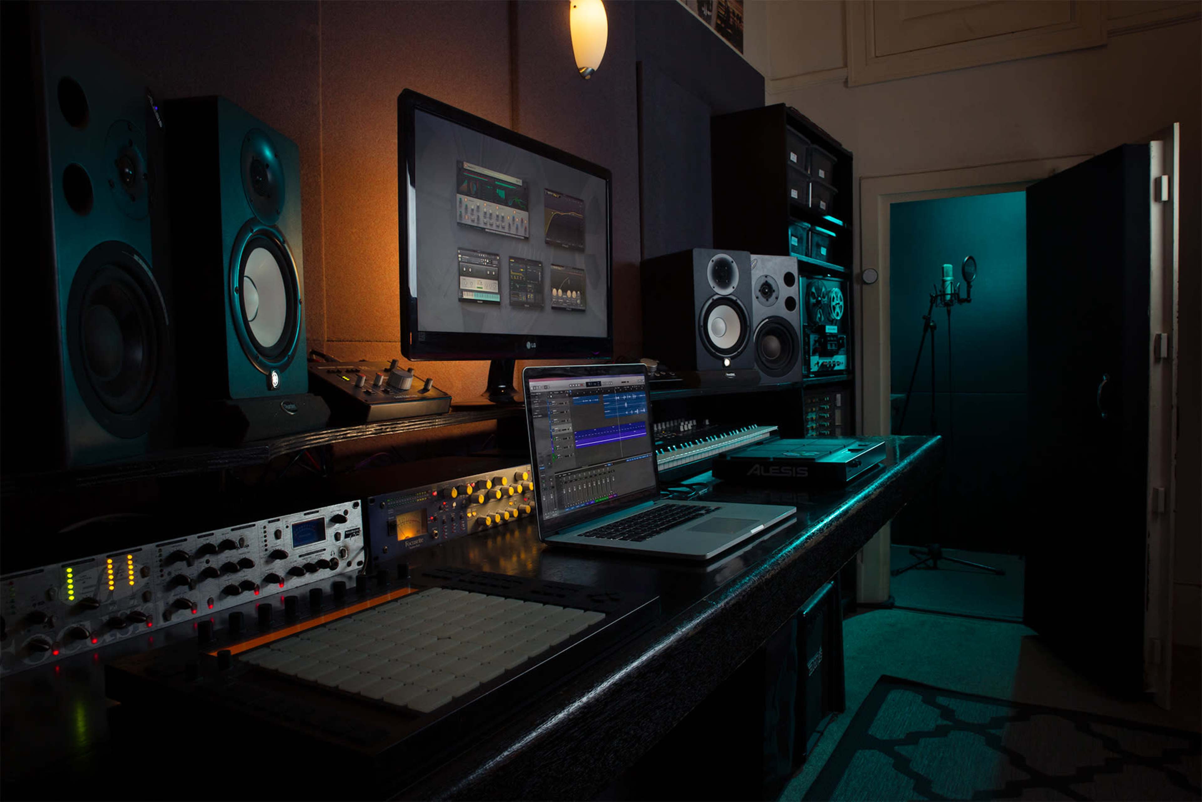 A music production studio contains a computer, audio interface, speakers, and a microphone set up in a dimly lit space.