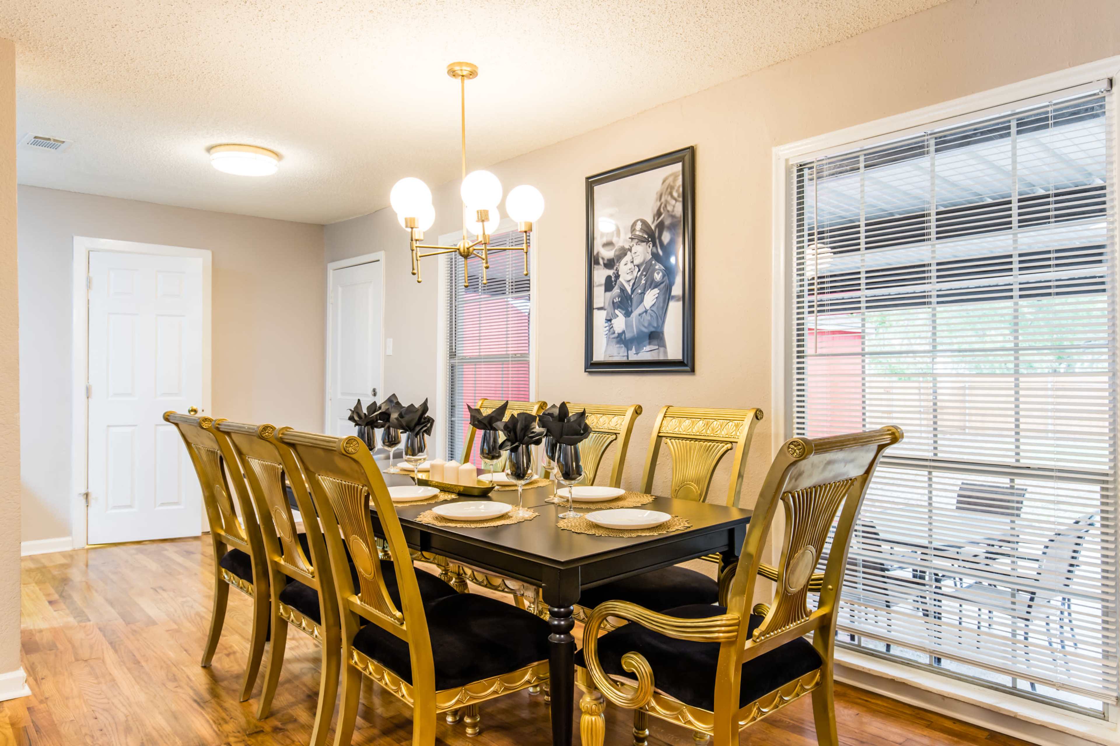 A large dining room features an elegantly set table surrounded by ornate gold chairs, with a framed black-and-white photograph on the wall and window blinds letting in natural light.