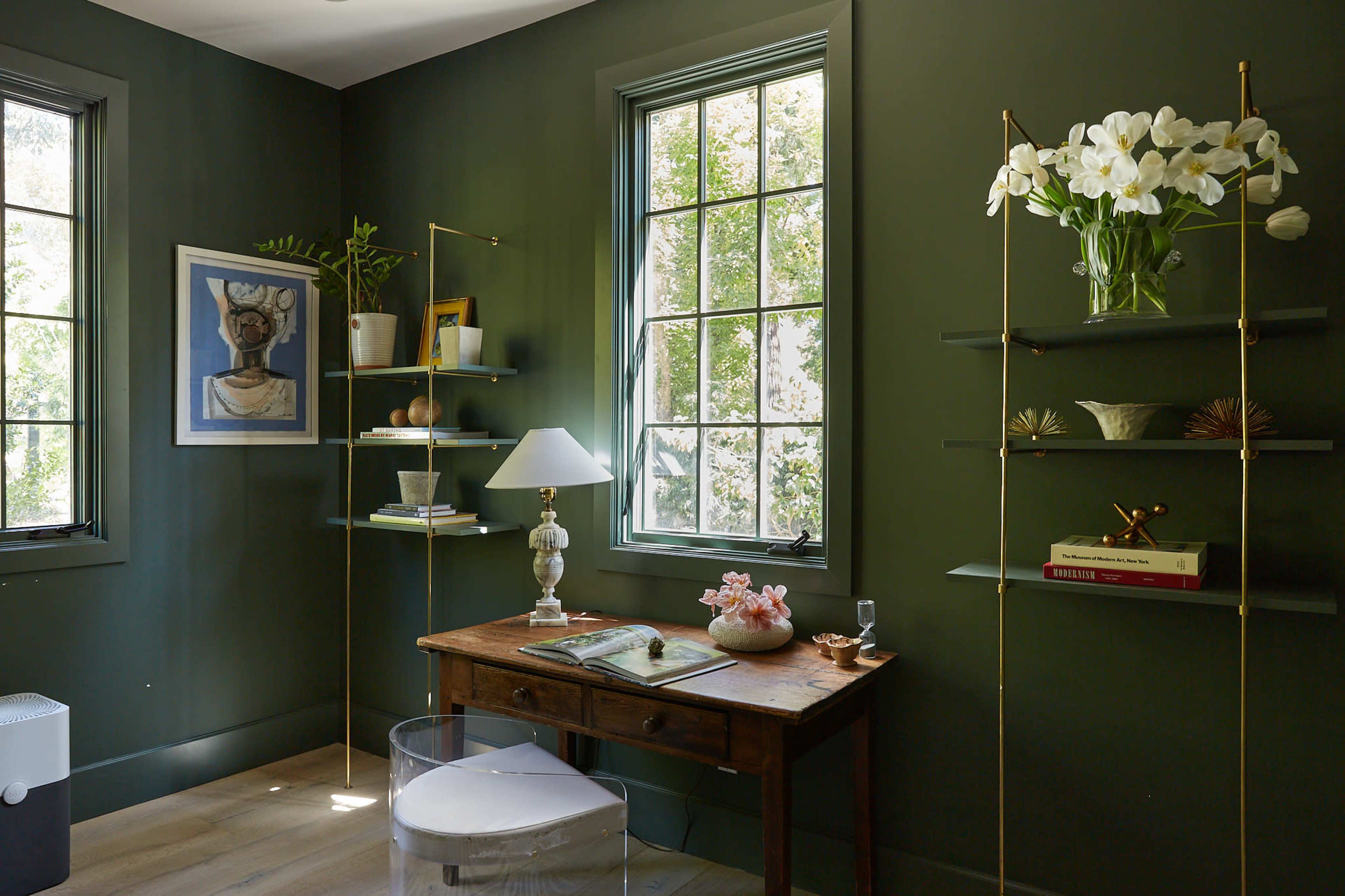 A small home office features a wooden desk with a clear acrylic chair, surrounded by green walls, large windows, and shelves displaying decorative items and plants.