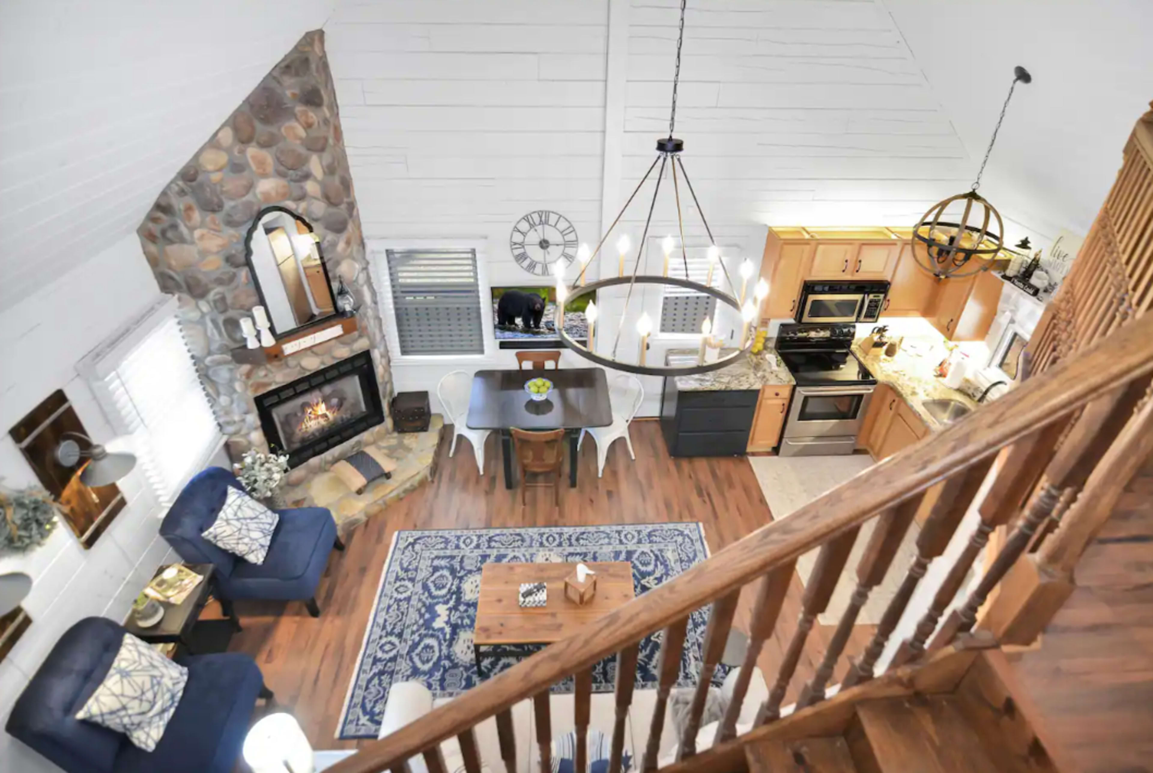 A spacious living area with a stone fireplace, a wooden dining table, and a well-equipped kitchen, all under a high ceiling with a modern chandelier.