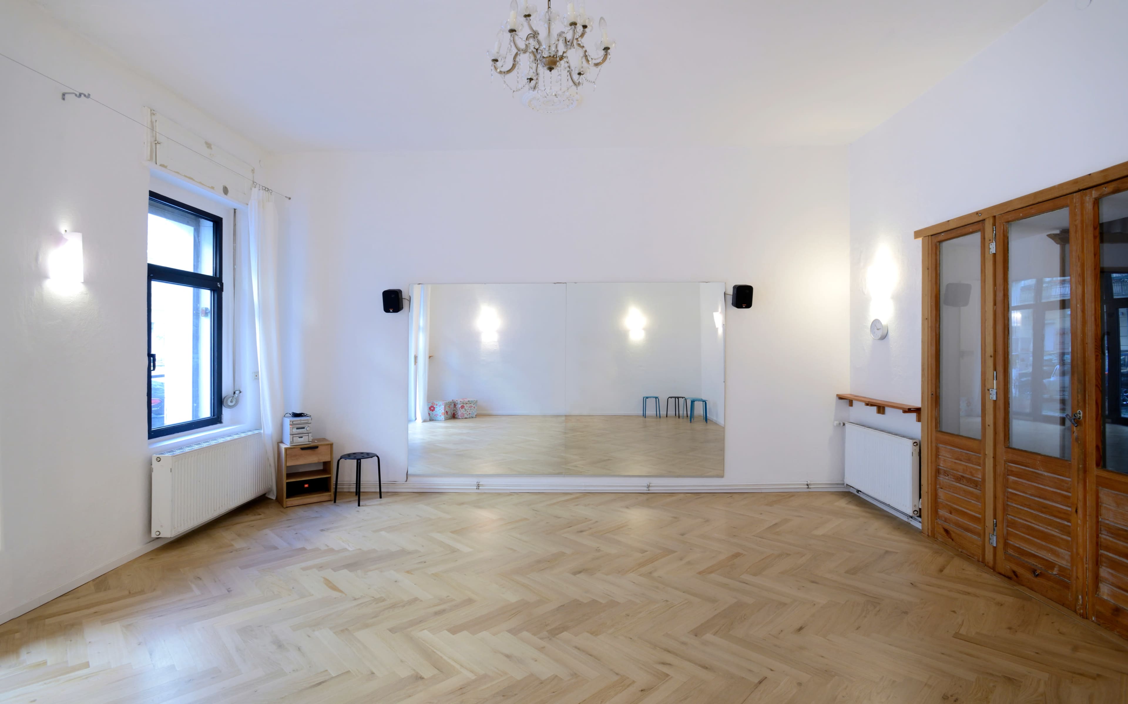 A spacious, empty room with a large mirror on one wall, wooden flooring, and a set of wooden double doors.