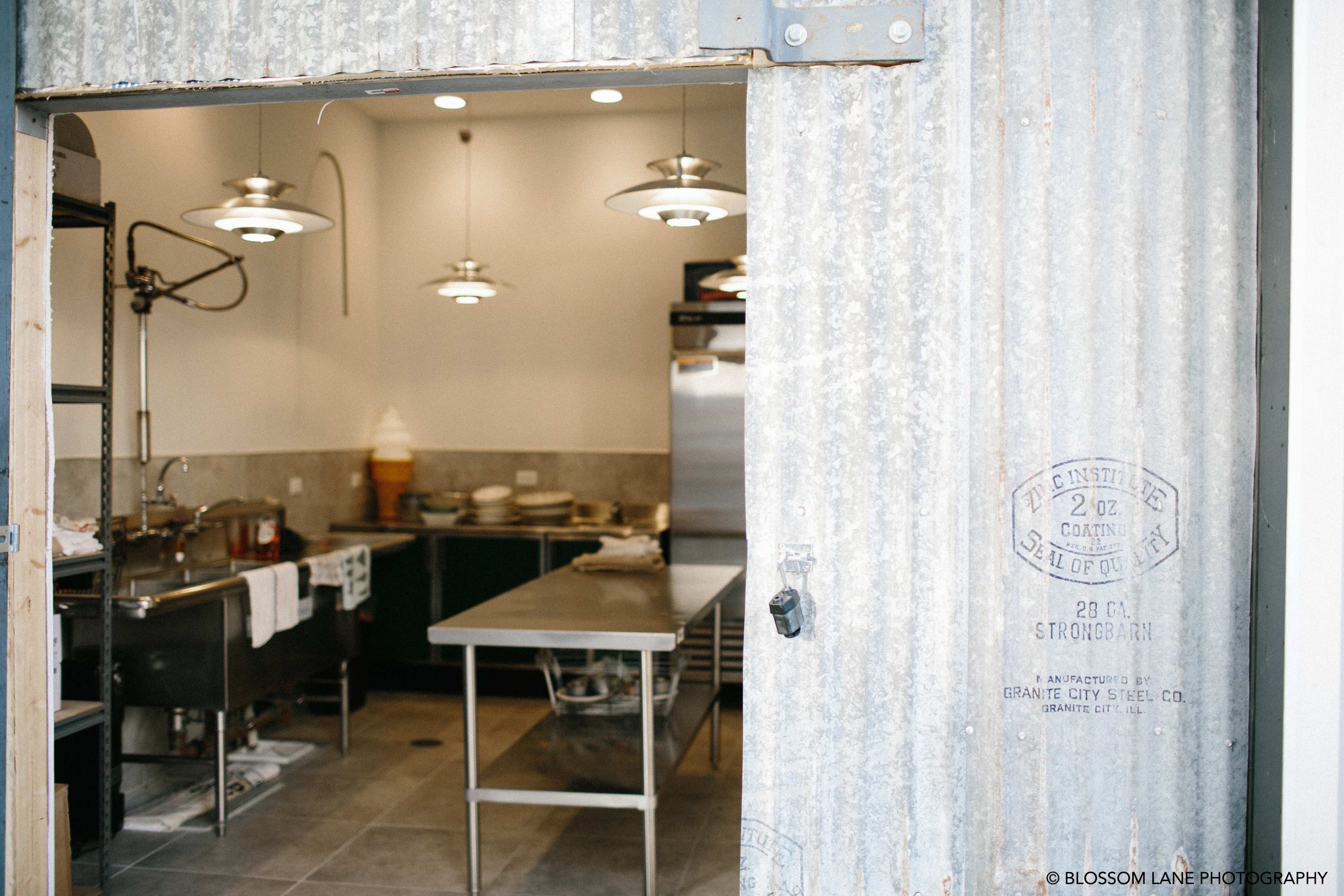 A commercial kitchen with stainless steel equipment and counters visible through a corrugated metal doorframe.