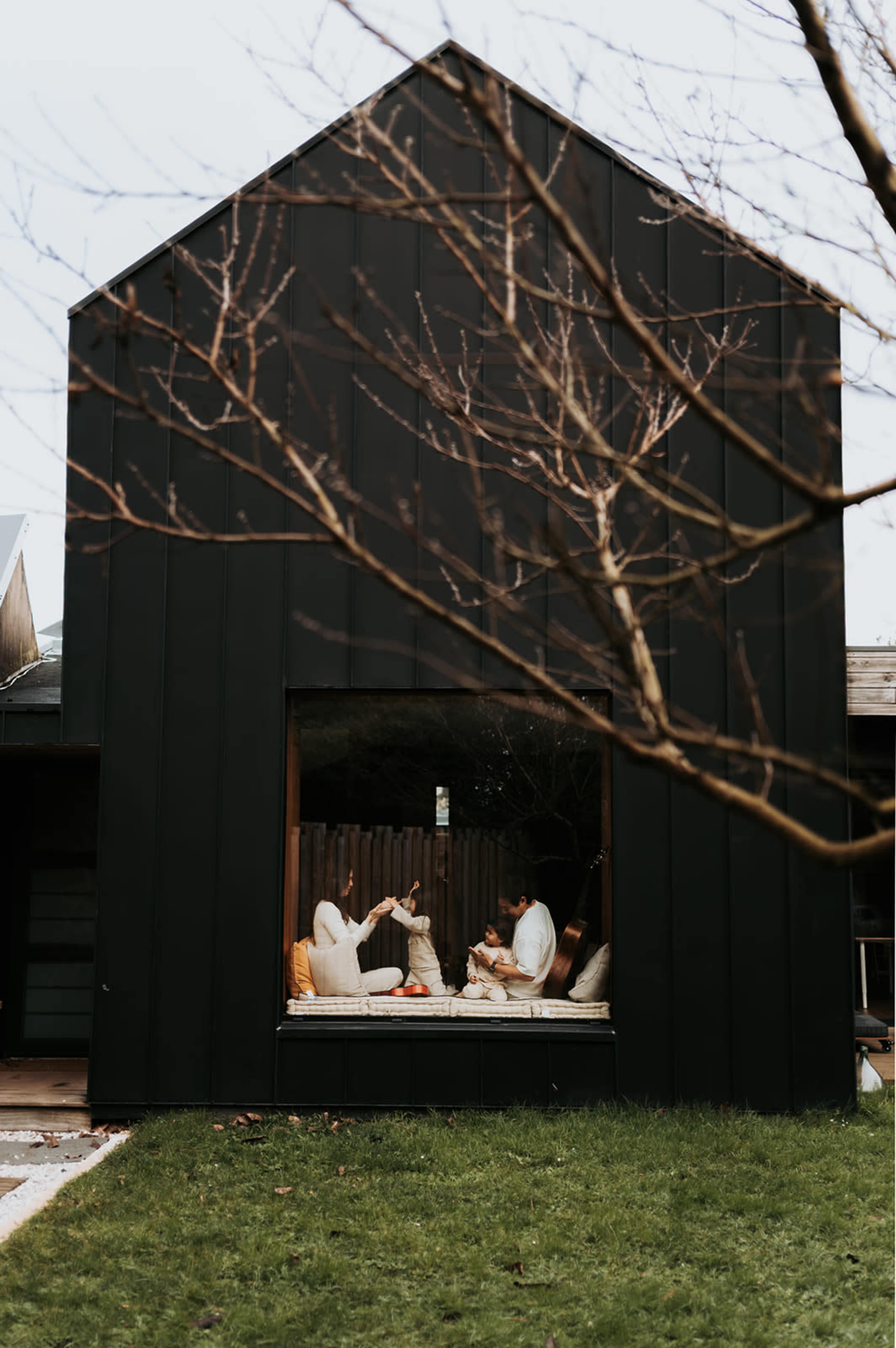 A family sits together on a built-in window seat inside a modern black house, surrounded by greenery and bare tree branches.