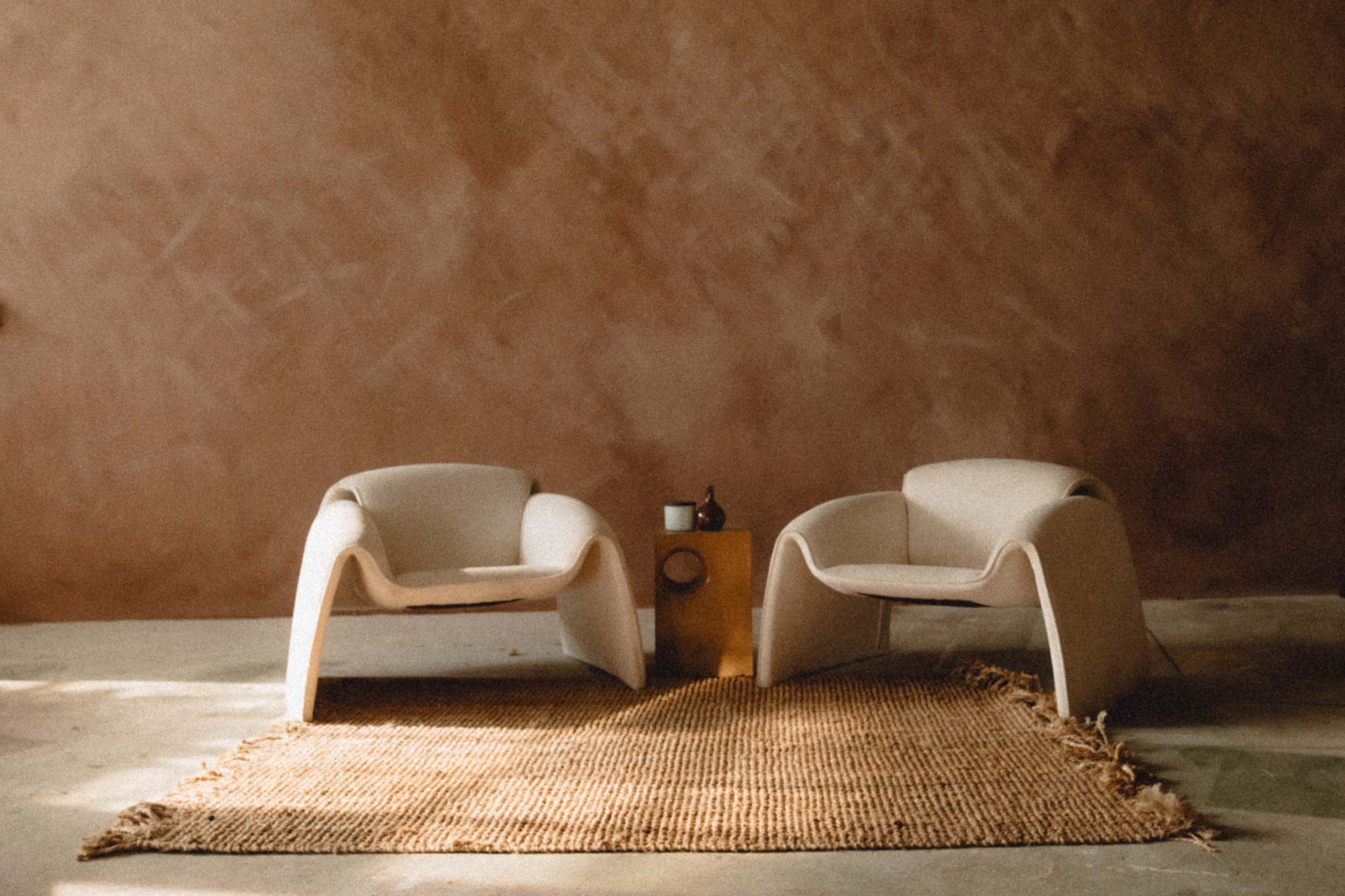 Two minimalist chairs are positioned on a woven rug in front of a small wooden side table against a textured brown wall.