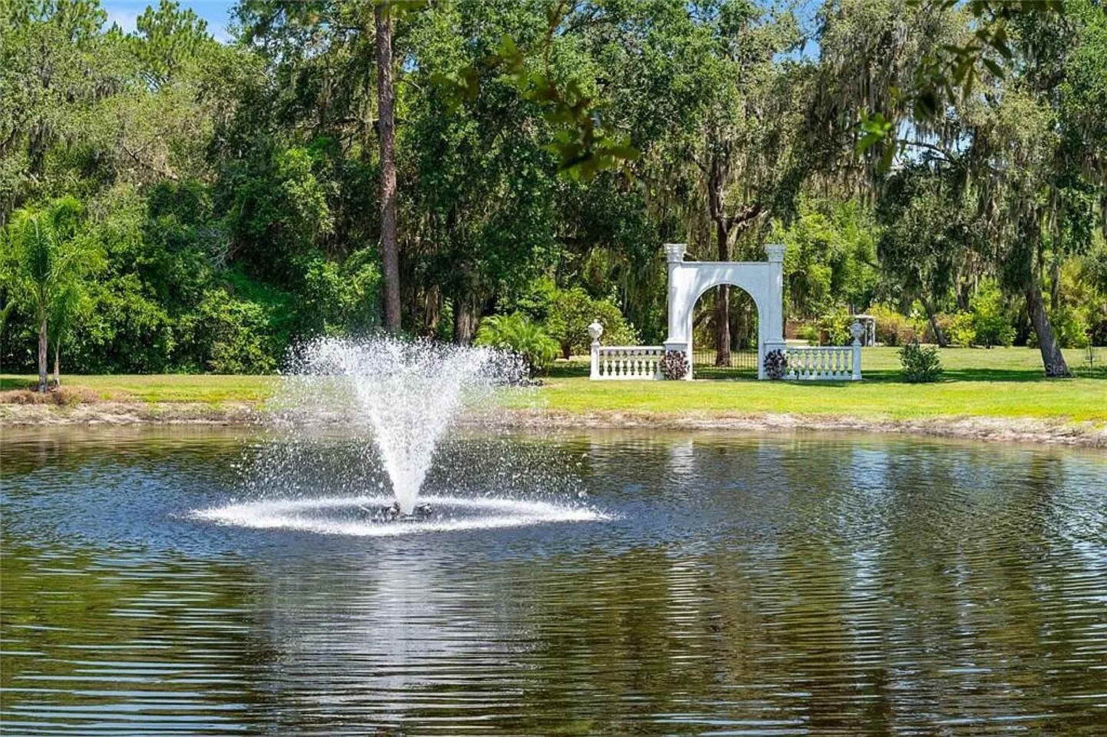 A fountain sprays water in a pond surrounded by lush greenery and a decorative white archway in the background.