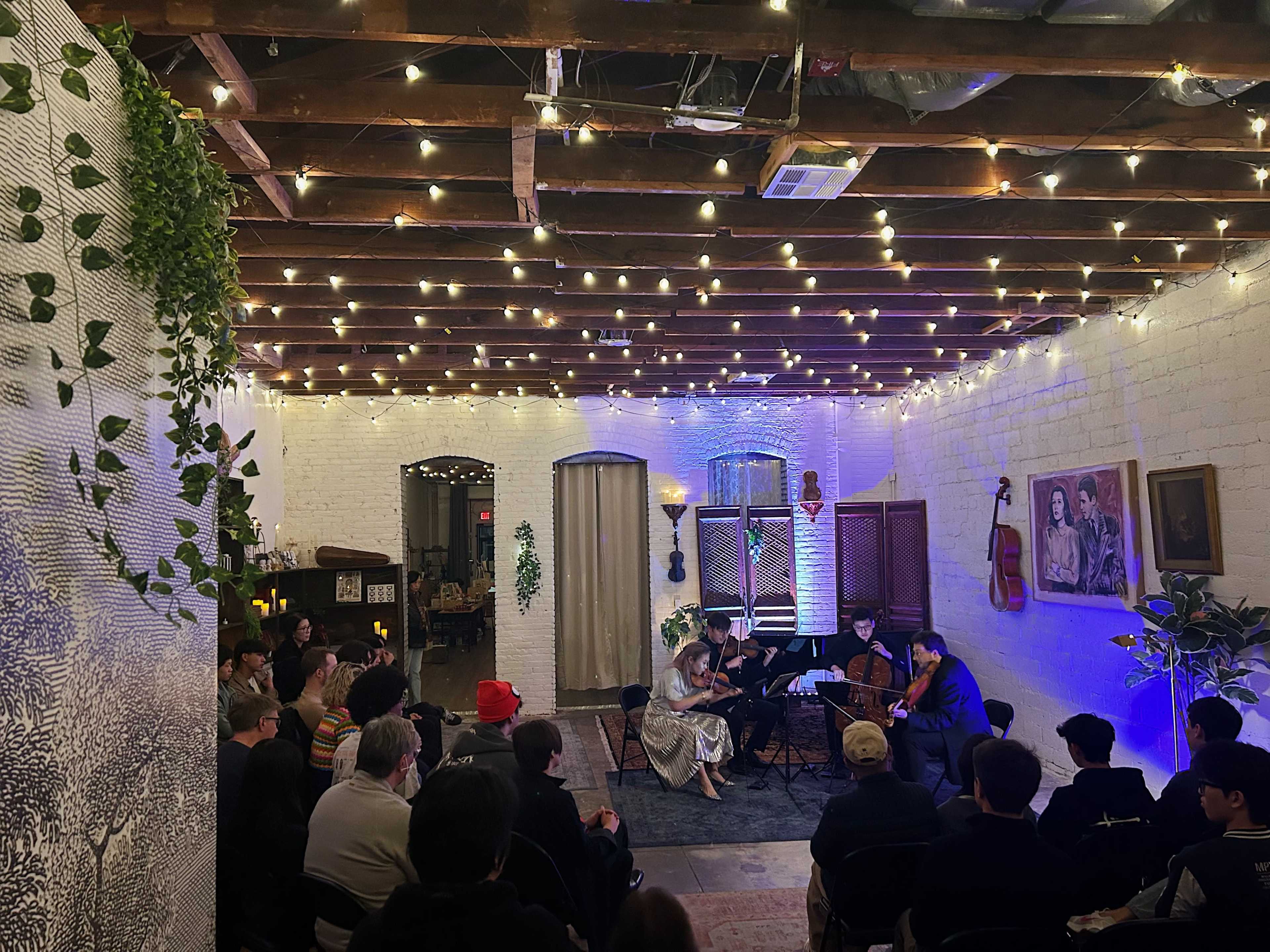 A small audience watches a live musical performance in a cozy, brightly lit venue with exposed beams and decorative plants.