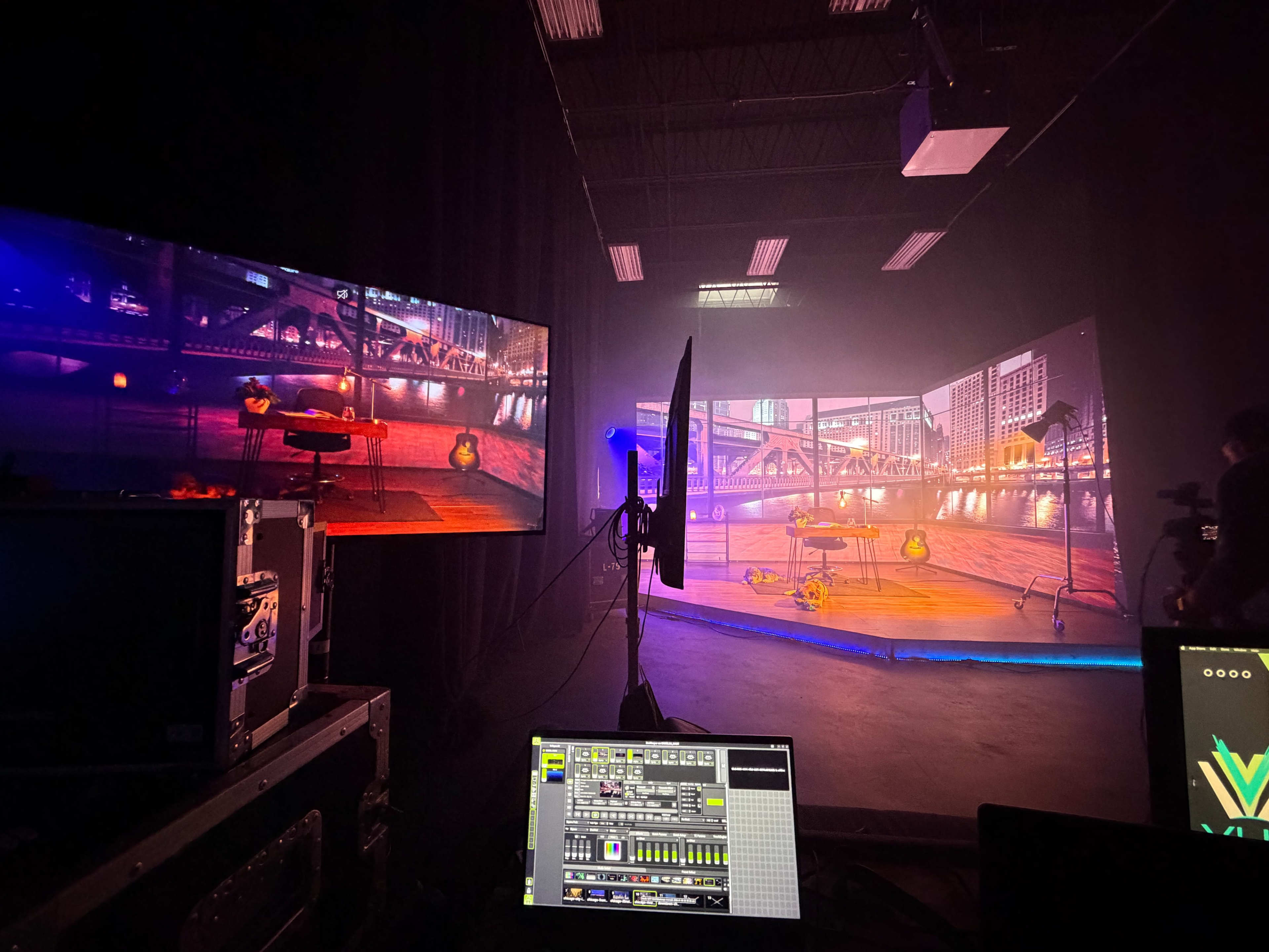 The image shows a dimly lit studio setup with large screens displaying a cityscape and equipment on a table, surrounded by a misty atmosphere.