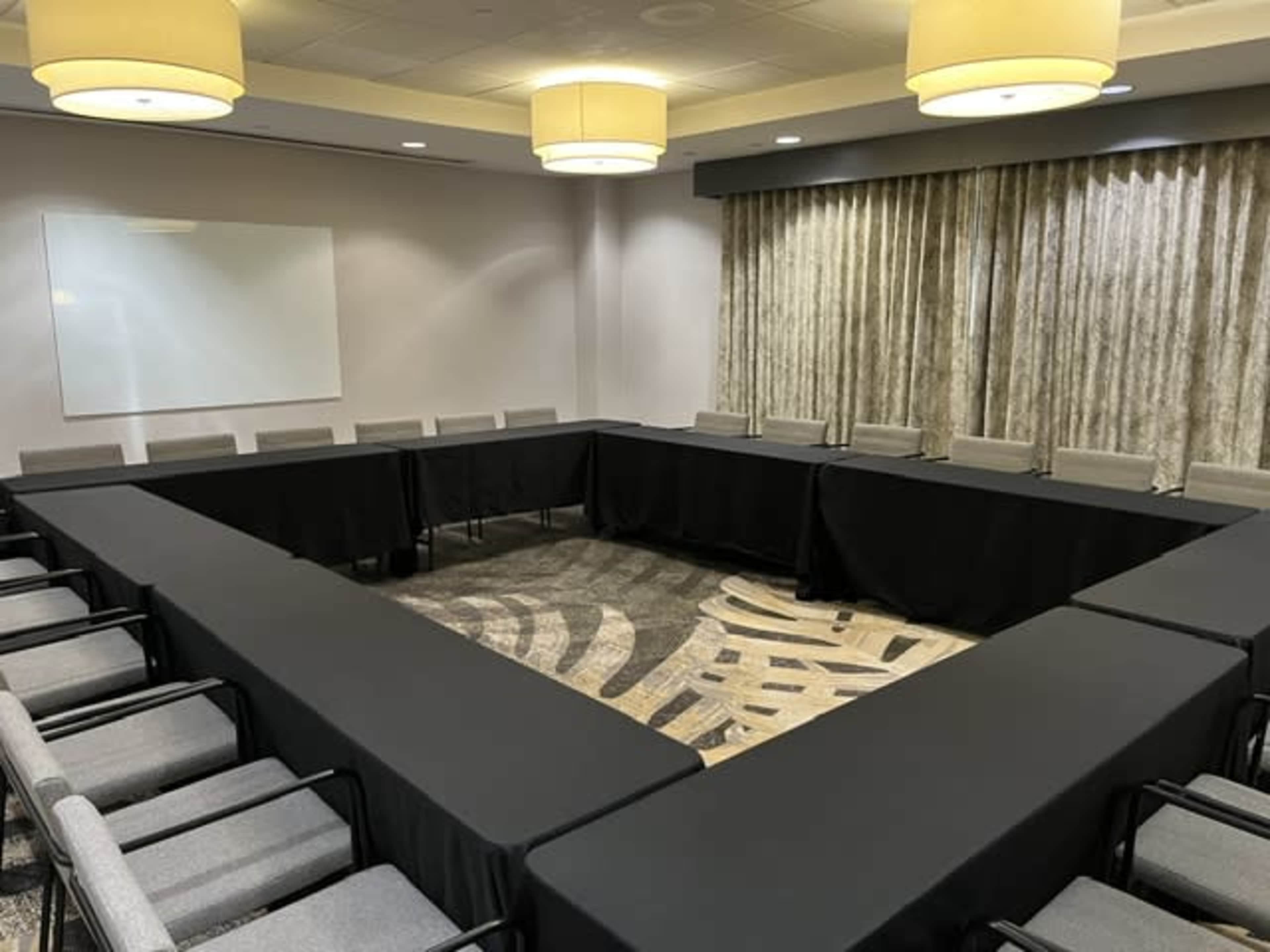 A meeting room features a large square arrangement of tables covered with black cloth and chairs around the perimeter, with soft lighting from overhead fixtures.