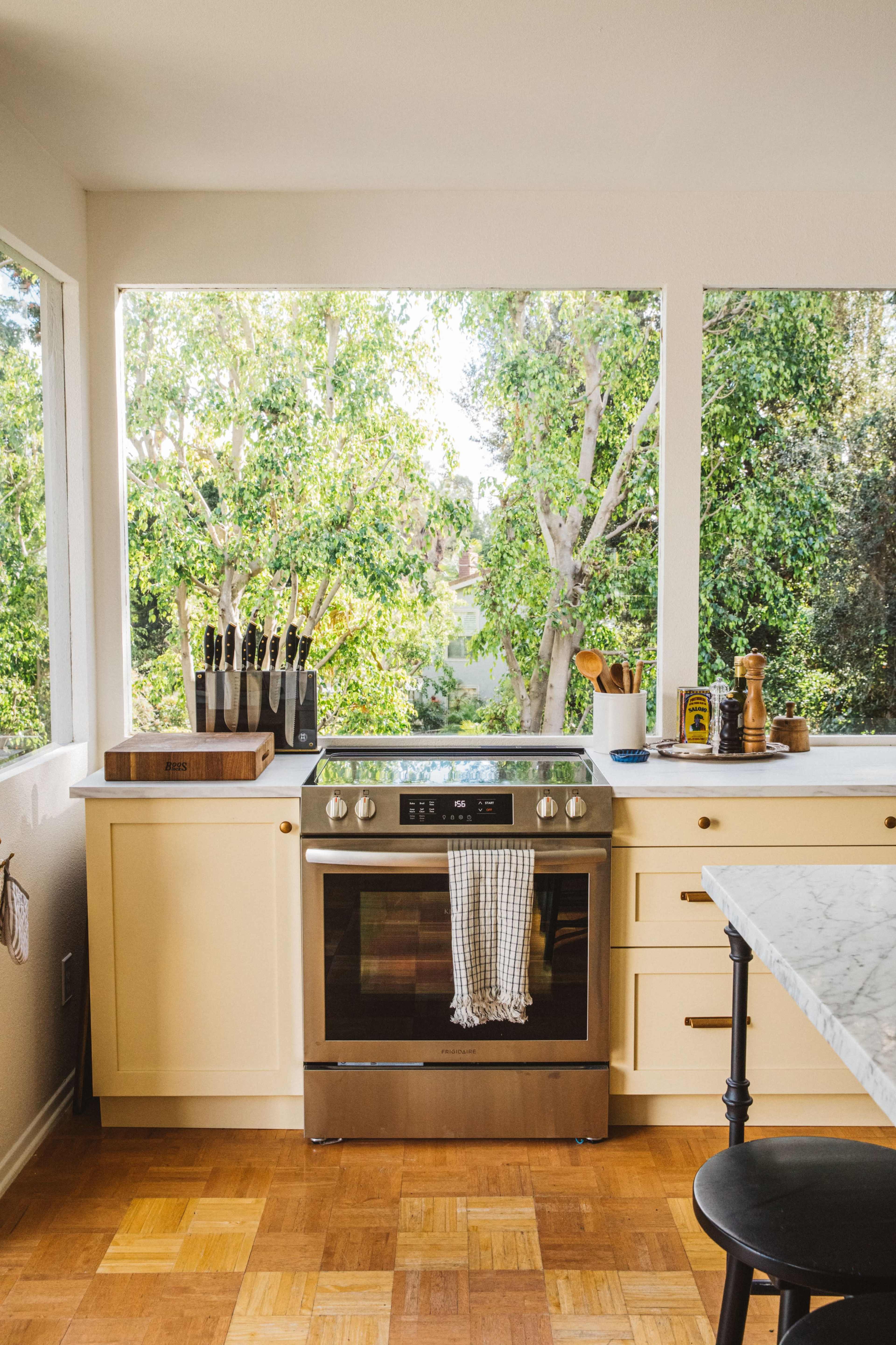 The kitchen features a stainless steel stove, a wooden knife block, and large windows that offer a view of the green landscape outside.