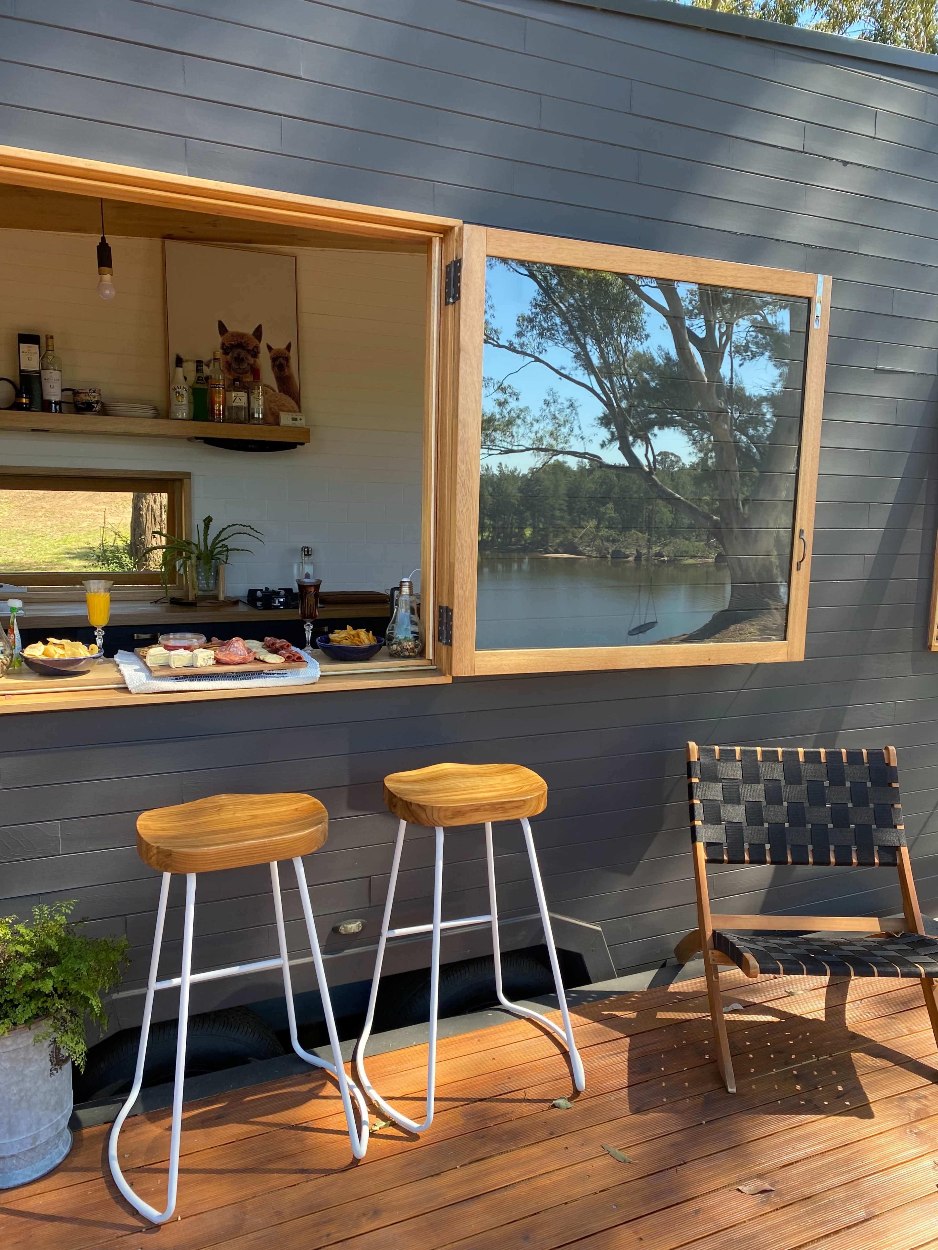 A wooden deck features two bar stools near an open window displaying a snack table and a view of a lake.