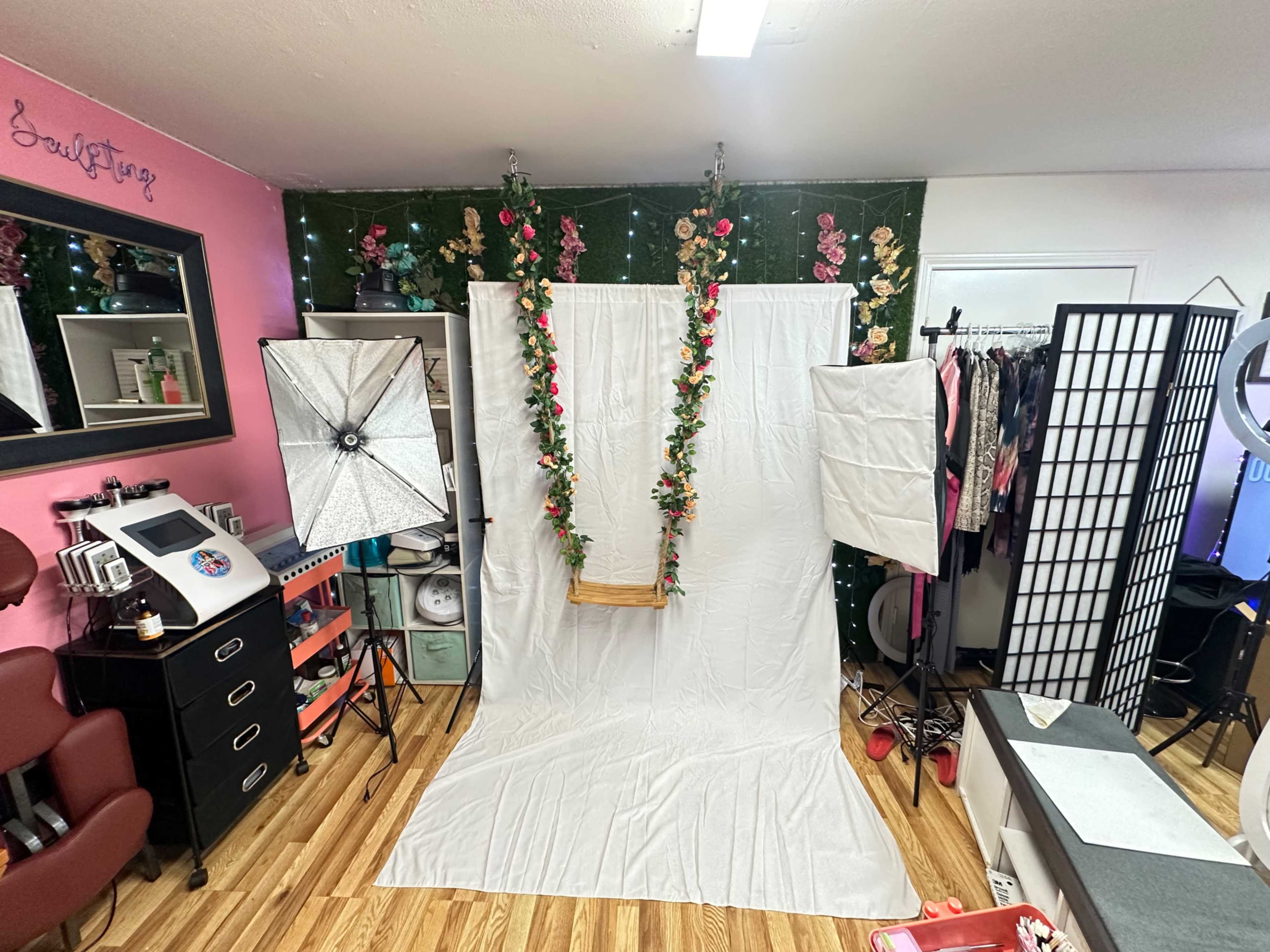 The image depicts a photography setup with a white backdrop, decorative flowers hanging from the ceiling, and two professional light stands positioned on either side.