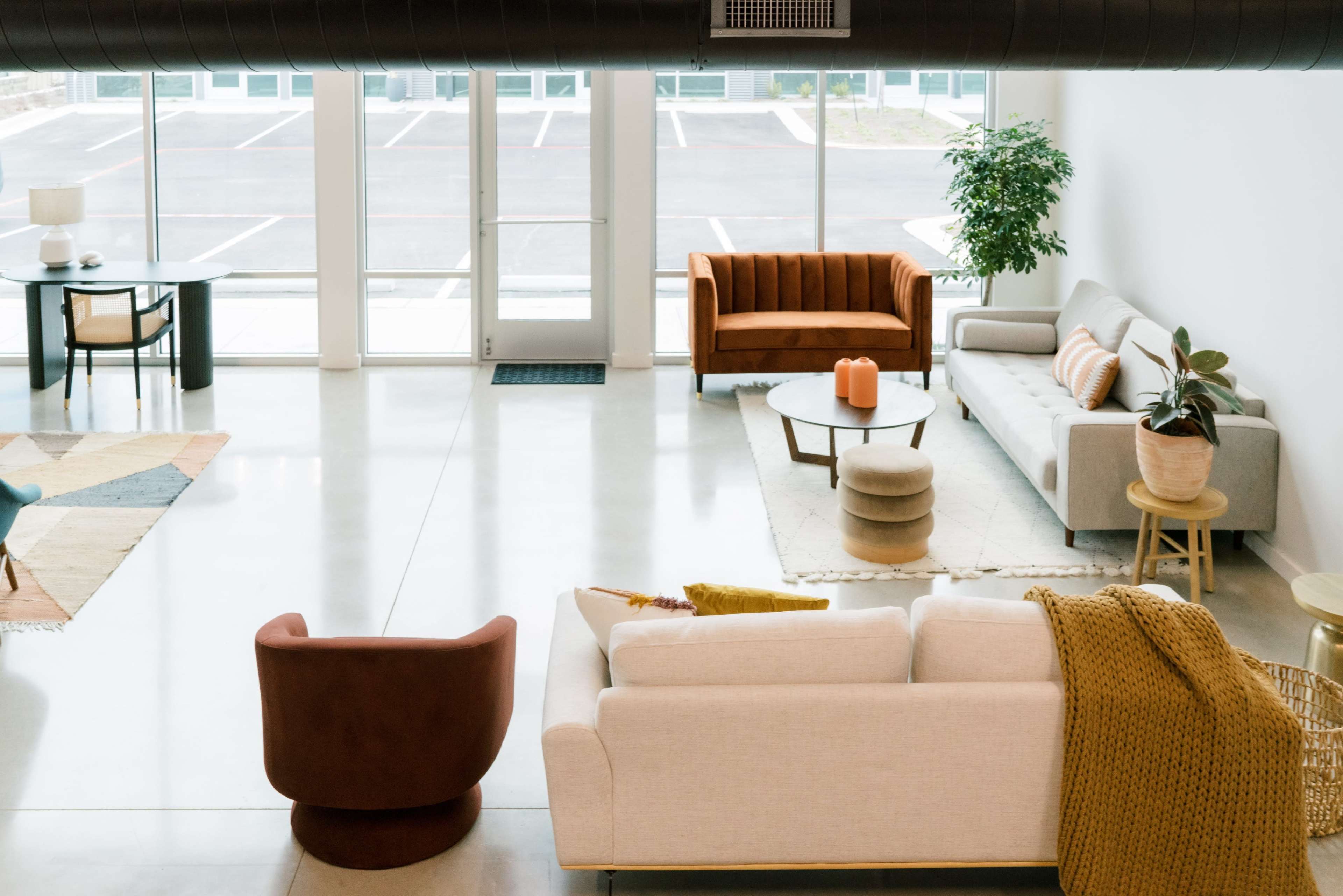 The image shows a spacious, modern living area furnished with two sofas, a coffee table, and a small dining table, all placed around a large window that lets in natural light.