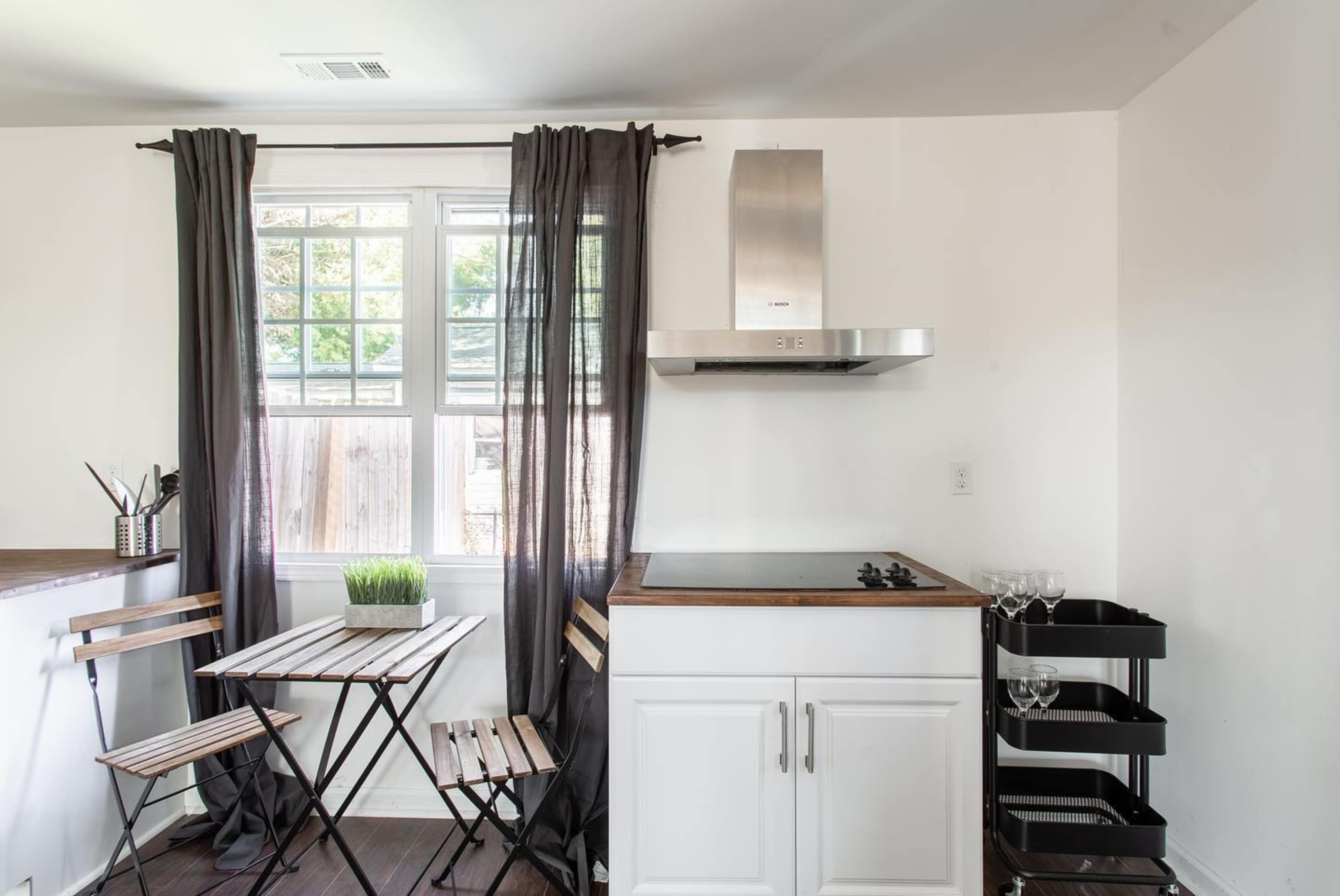 A small kitchen features a wooden table with two folding chairs, a stainless steel range hood above a cooktop, and a window dressed with black curtains.