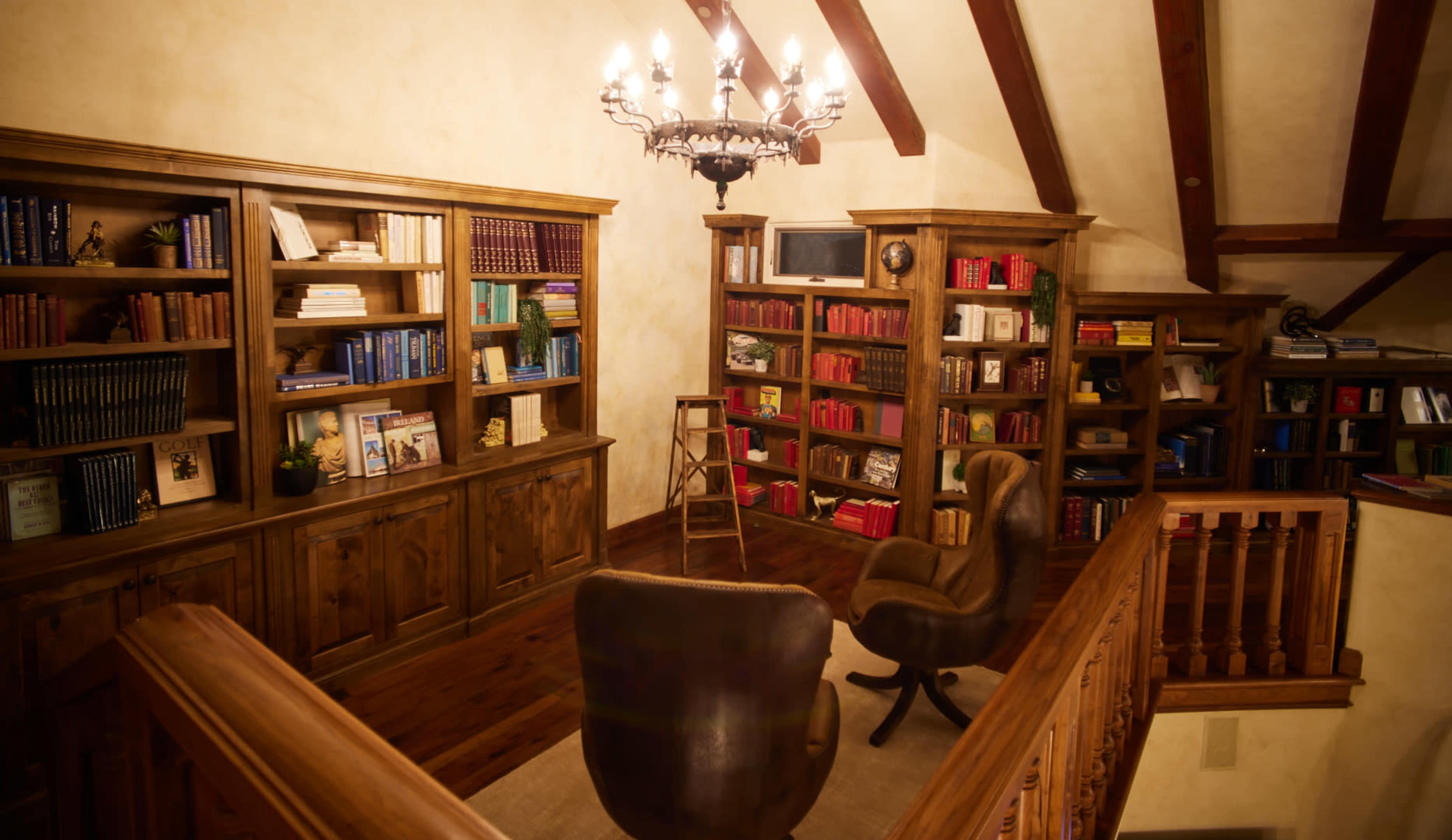 A cozy library with wooden bookshelves filled with books, a chandelier overhead, and two chairs positioned on a carpeted floor.