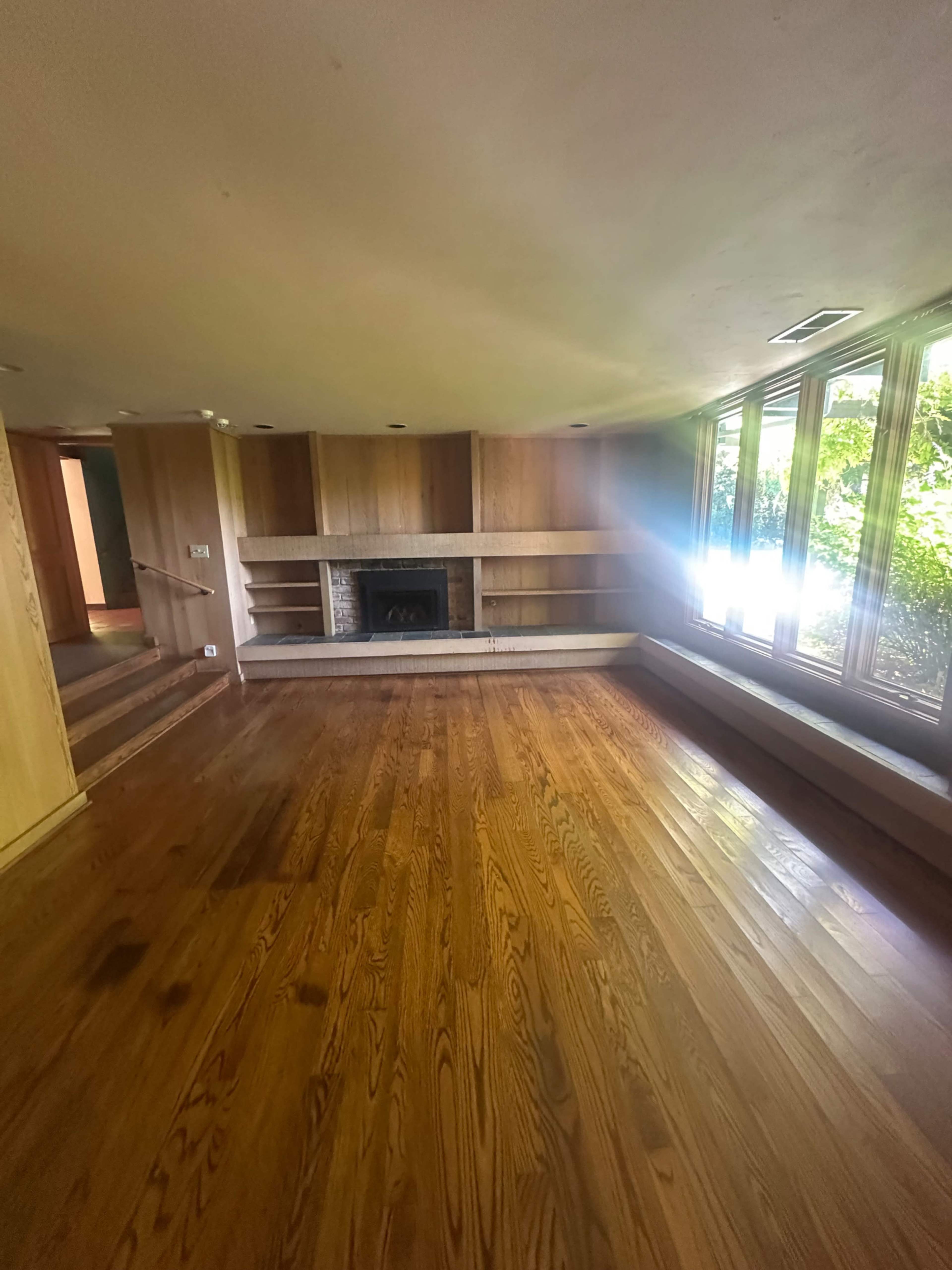 The image shows a spacious room with wooden flooring, a simple wall shelf, a fireplace, and large windows allowing natural light.
