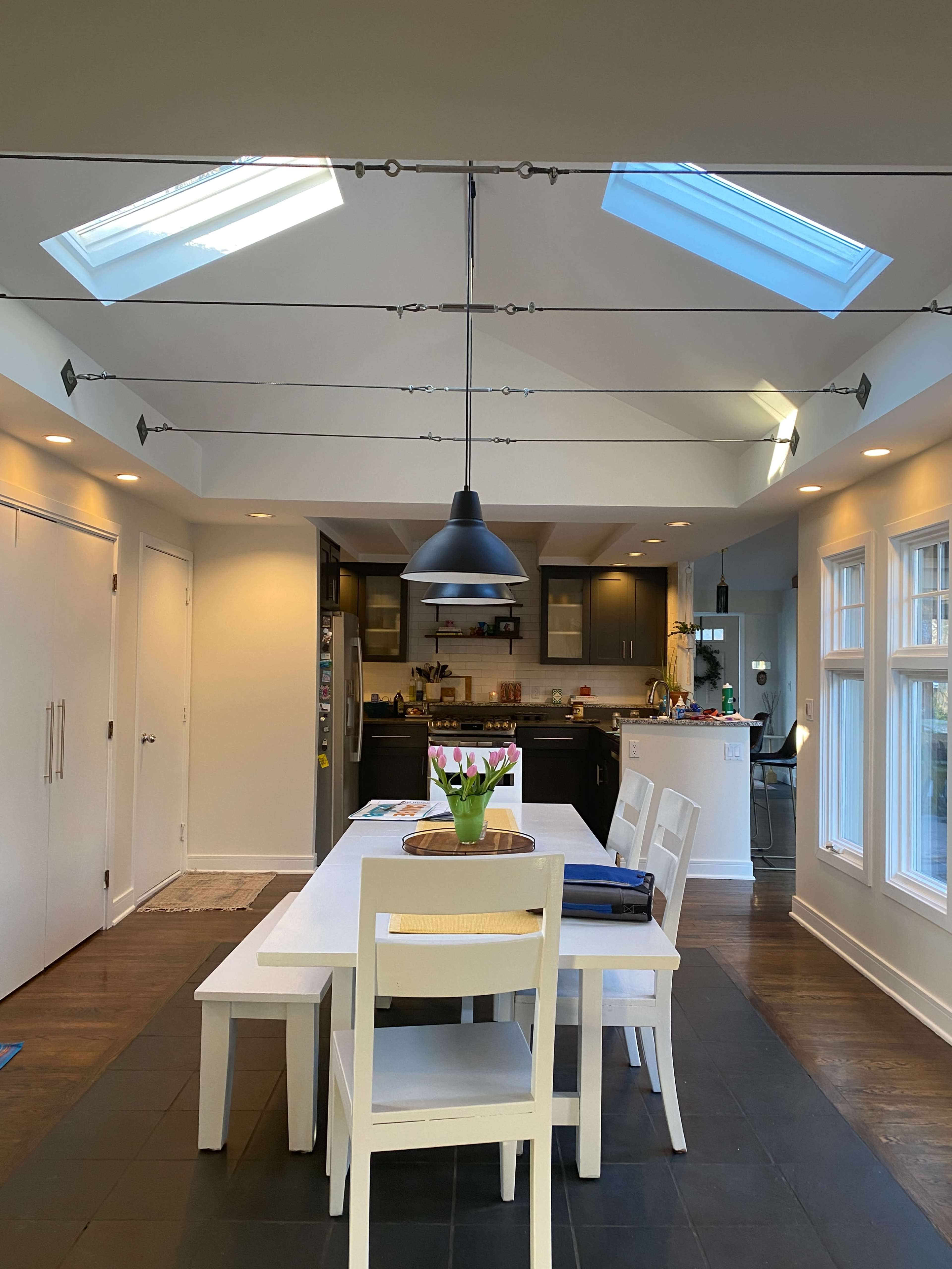 The image shows a modern kitchen and dining area featuring a white table surrounded by chairs, with skylights above and a dark-colored kitchen in the background.