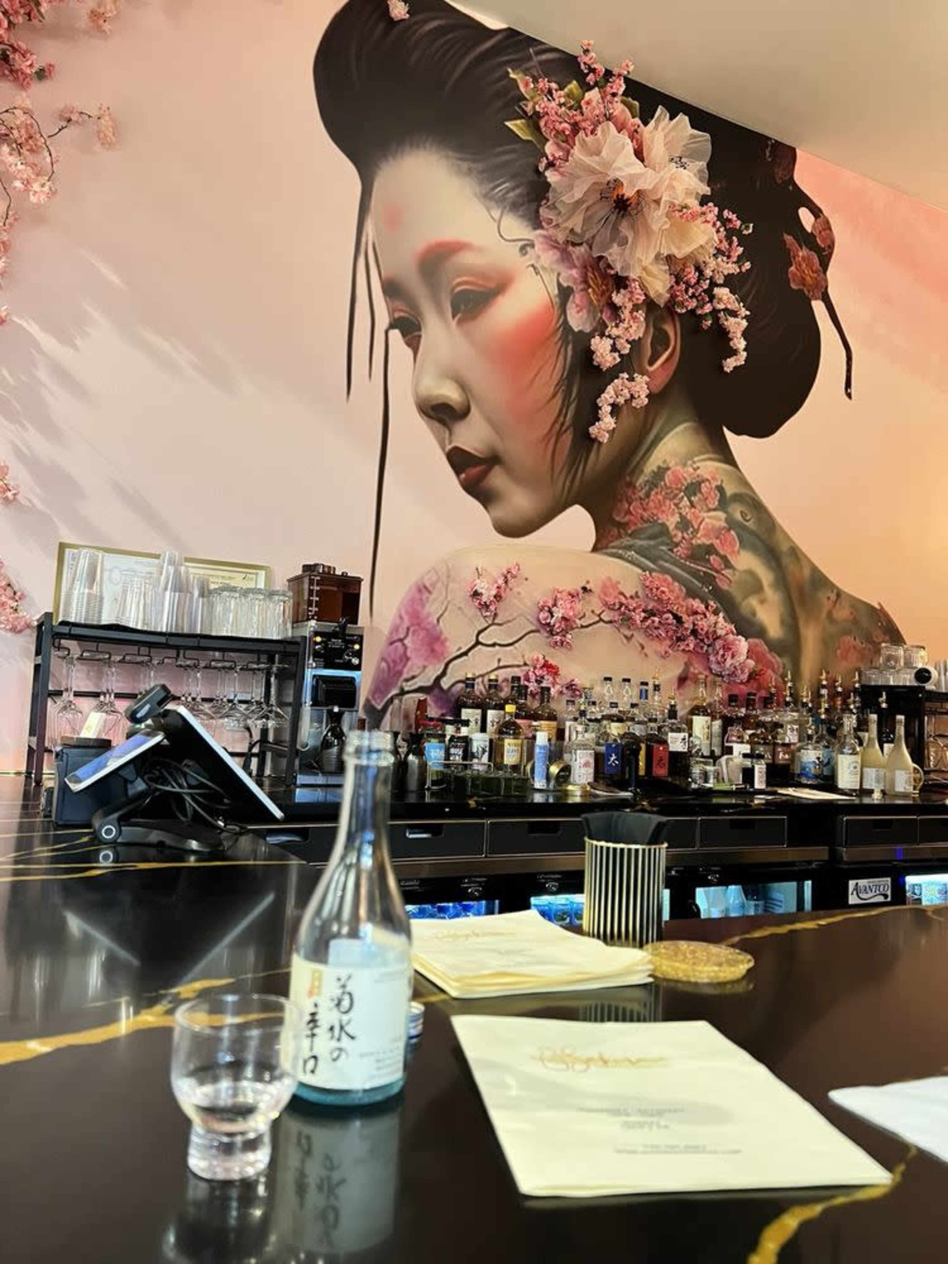The image shows a bar with a large mural of a woman adorned with flowers and traditional Japanese elements, alongside a sake bottle and a glass on the counter.