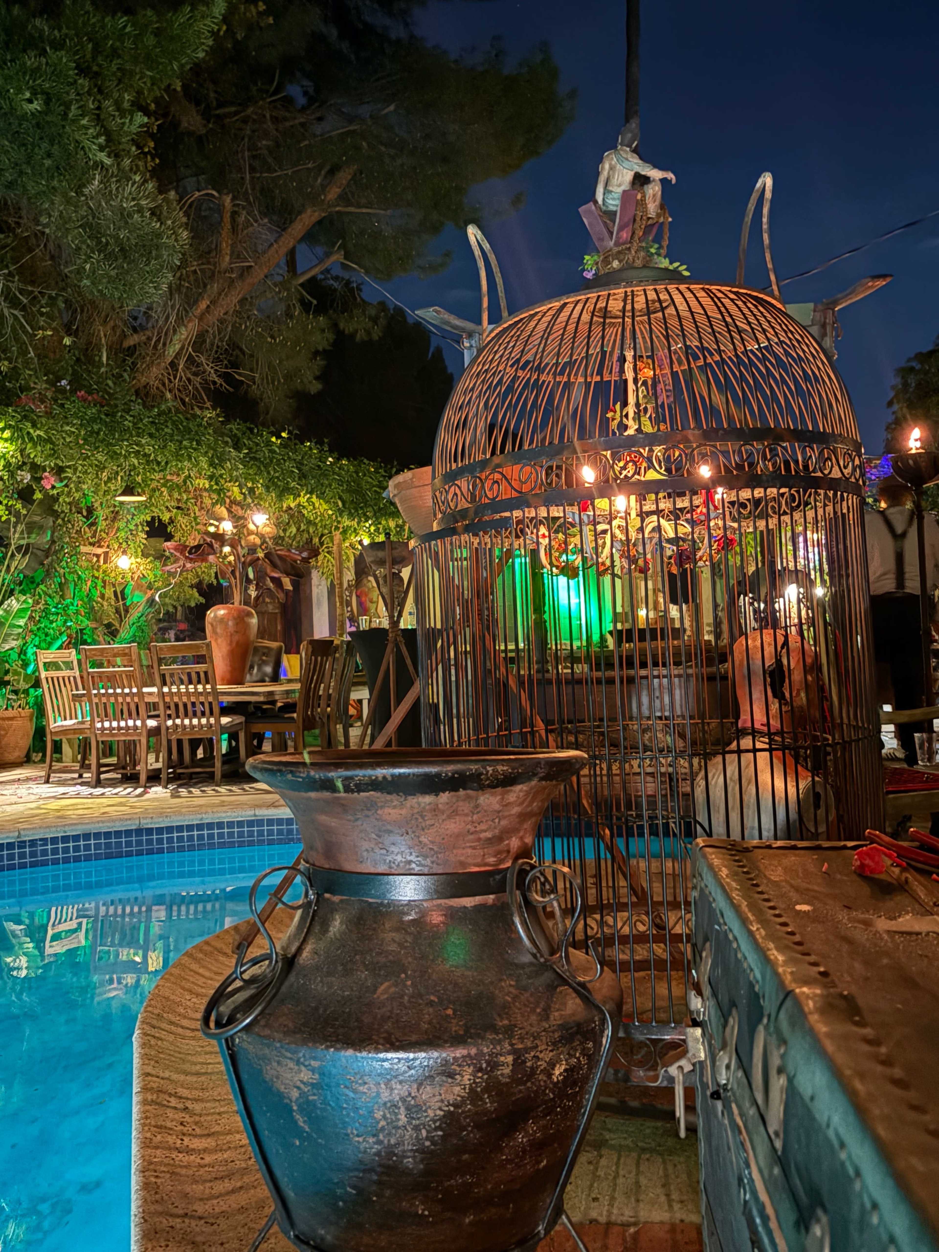 A large, decorative birdcage stands beside a pool, with various plants and outdoor furniture illuminated by warm lights in the evening.
