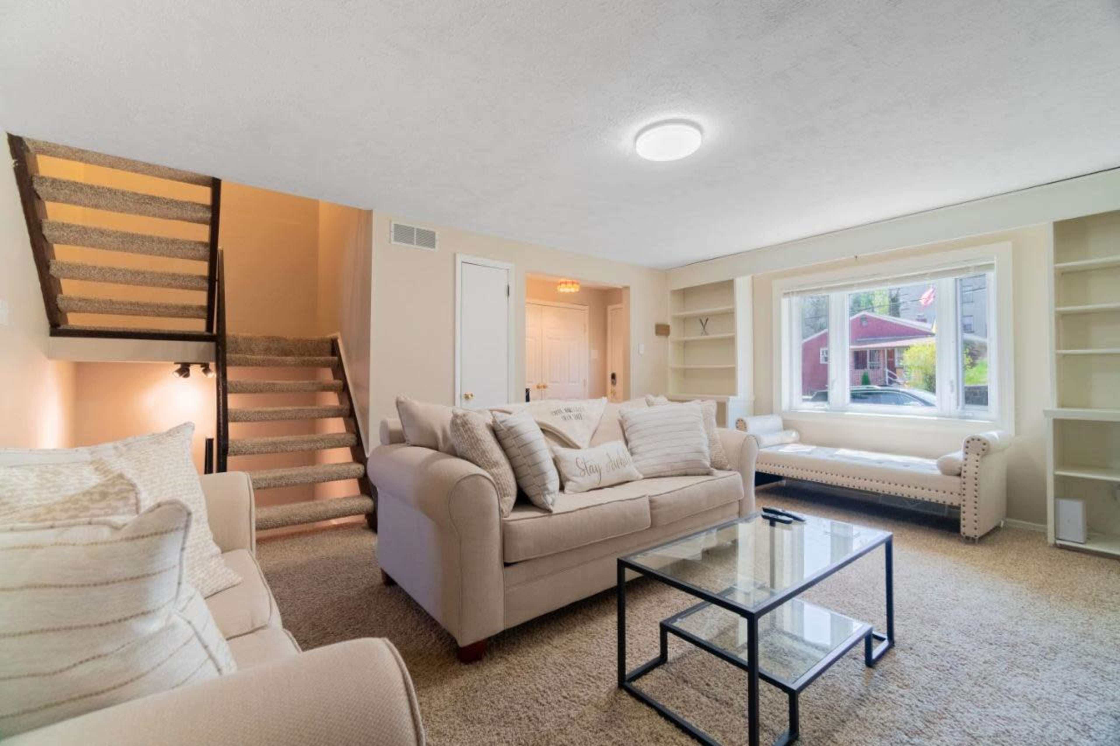 A bright living room features beige sofas, a glass coffee table, and a large window overlooking the outdoor space, with stairs leading to a second level.