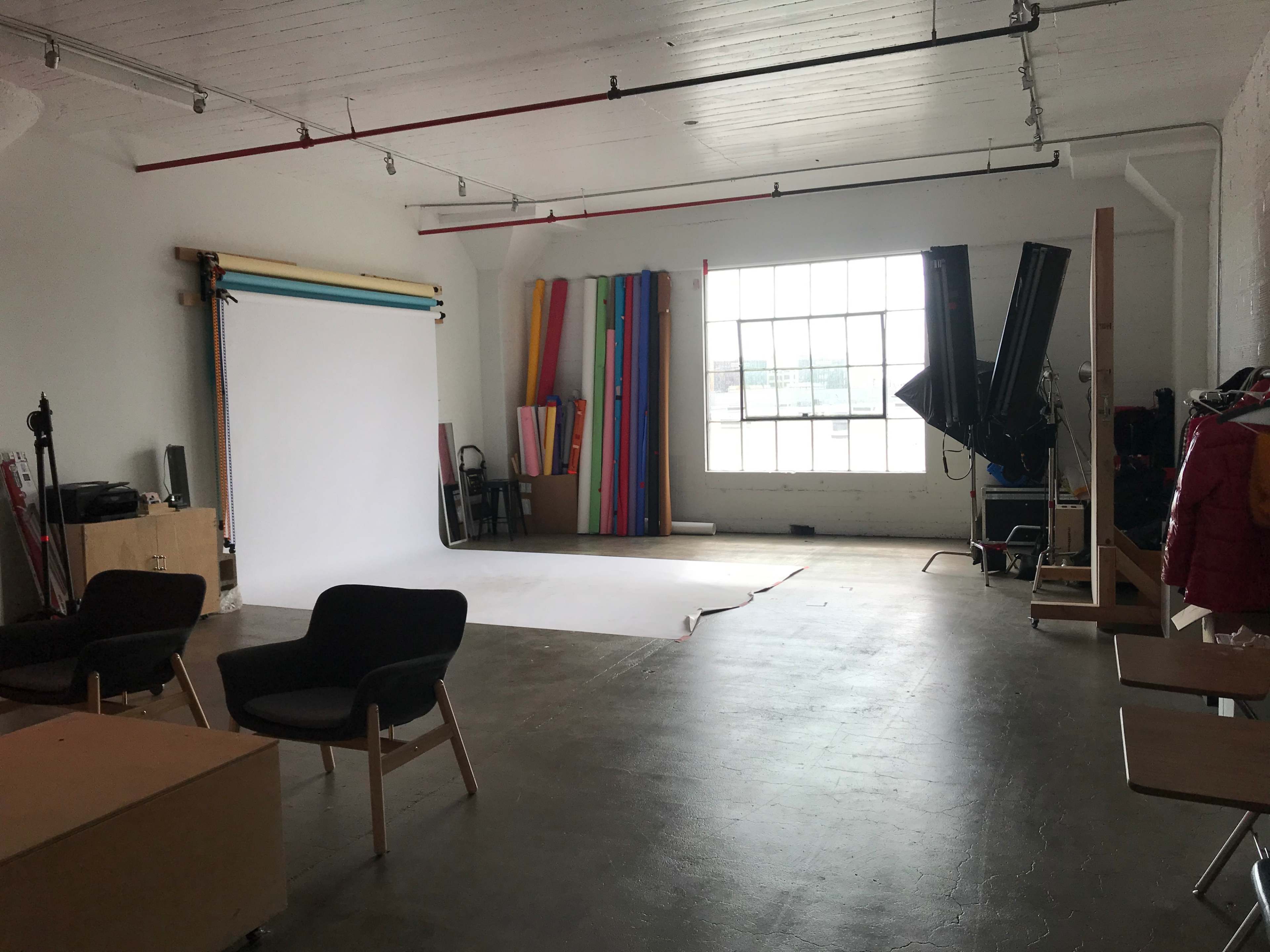 A spacious photo studio features a white backdrop, colorful rolls of paper, and various equipment arranged near large windows.