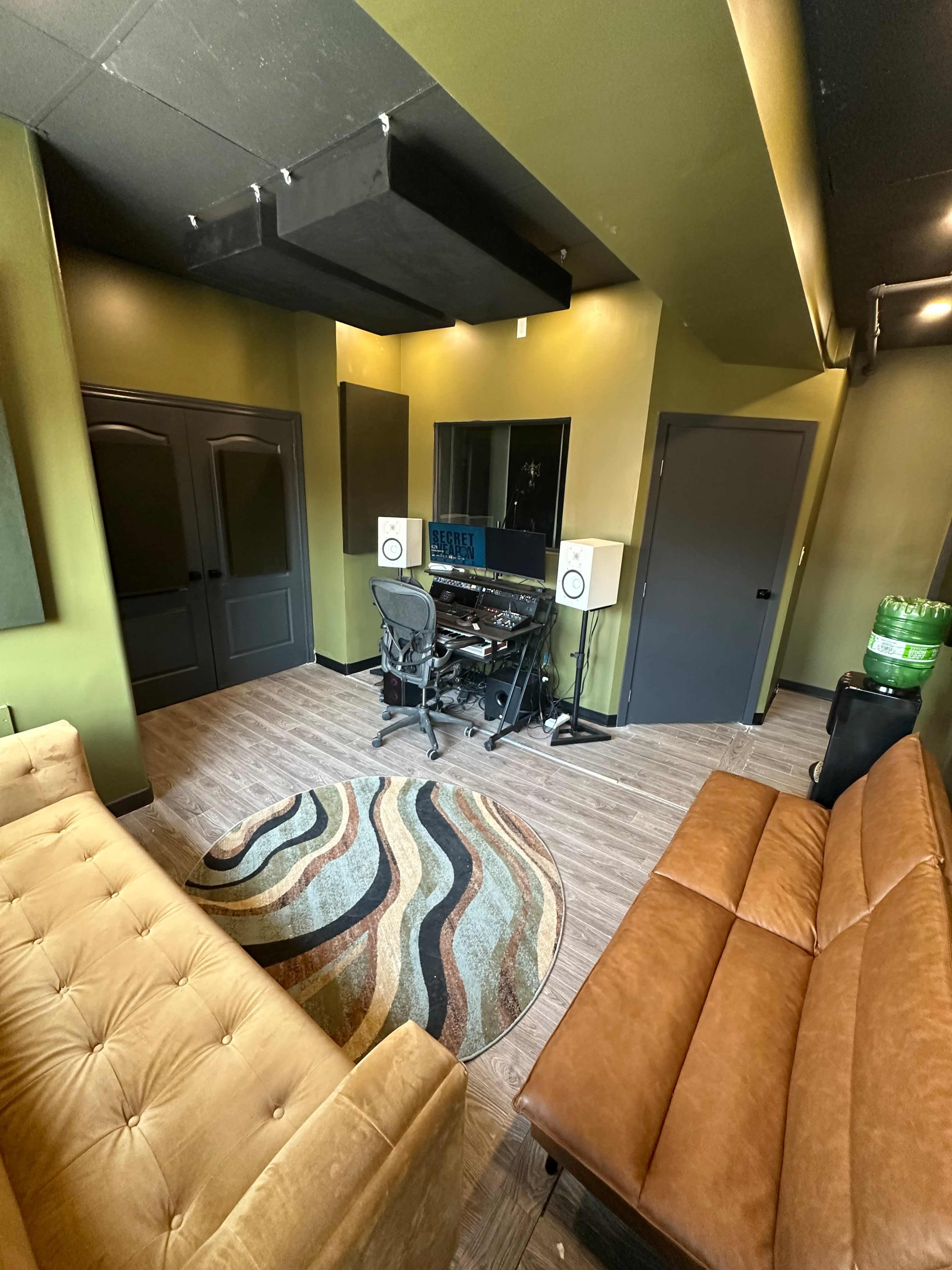 The image shows a recording studio with a desk, audio equipment, and a comfortable seating area featuring a brown sofa and a round rug on the floor.