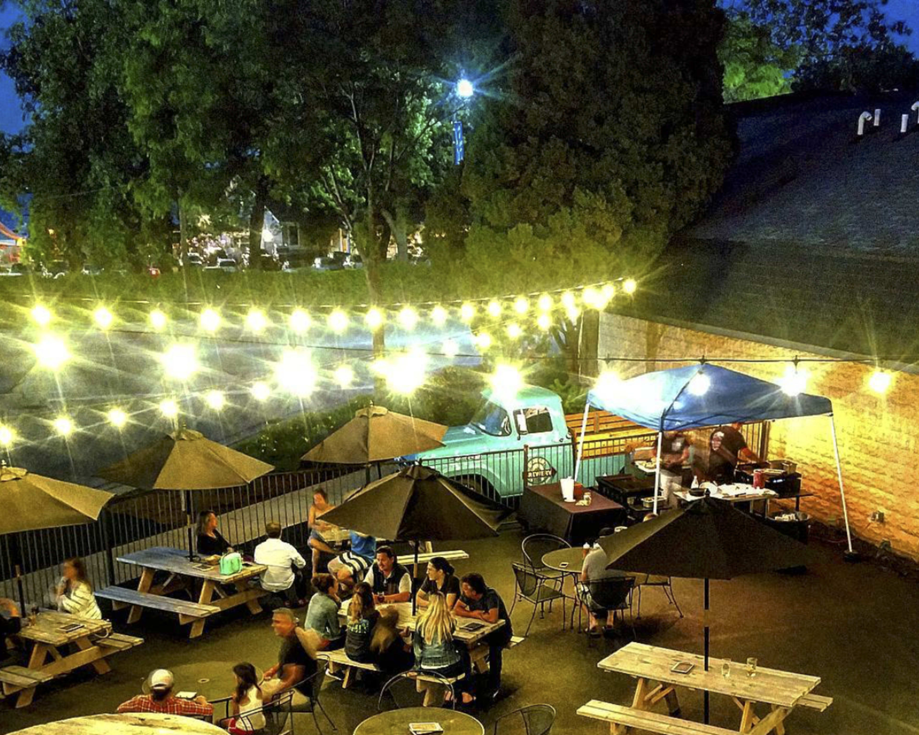 An outdoor dining area illuminated by string lights features numerous tables filled with people and a food stall in the background.