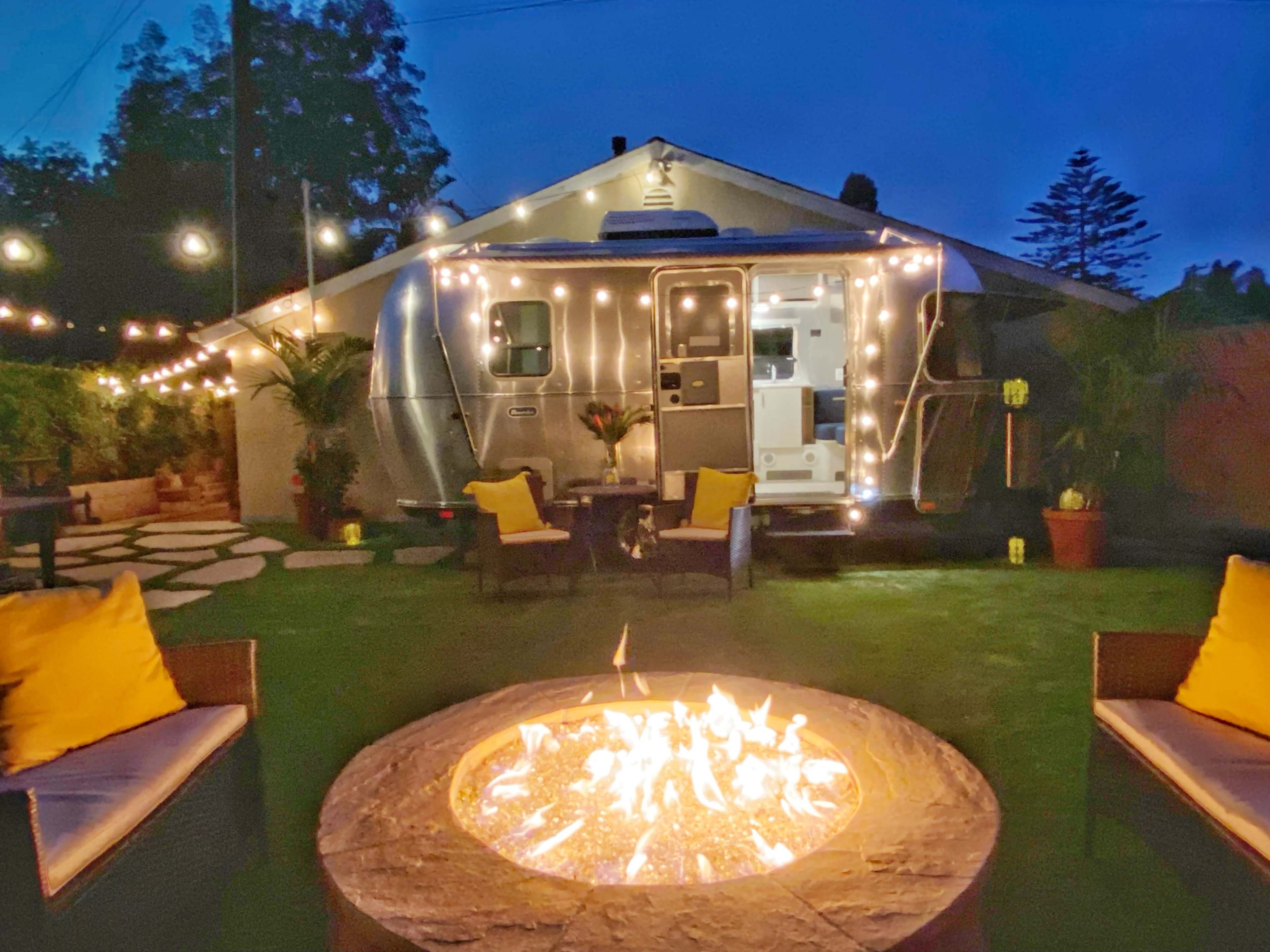 The scene features a backyard with an illuminated Airstream trailer and a fire pit surrounded by chairs, all set against a twilight sky.