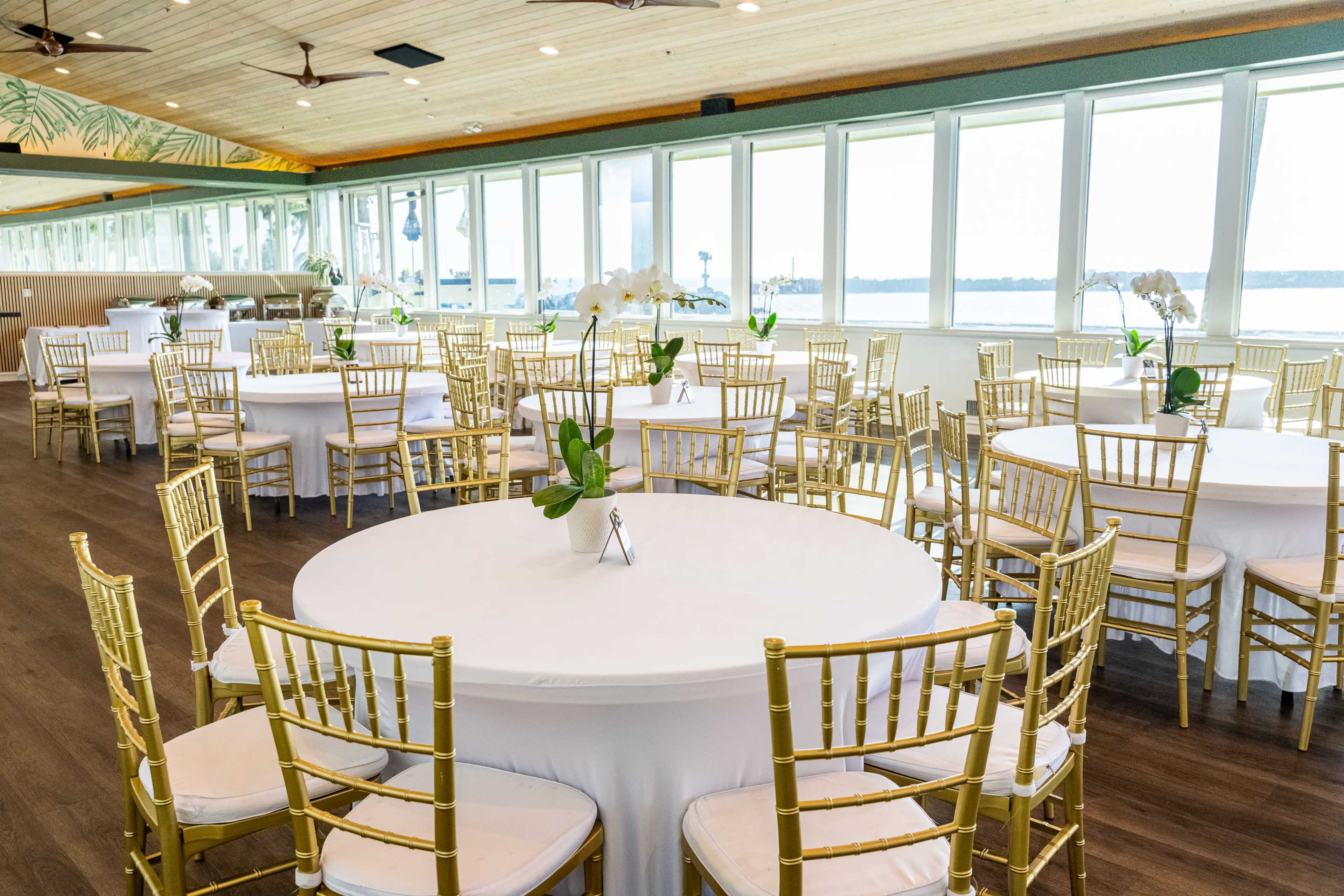 The image shows a bright event space with multiple round tables covered in white tablecloths, surrounded by gold chairs and large windows overlooking a body of water.