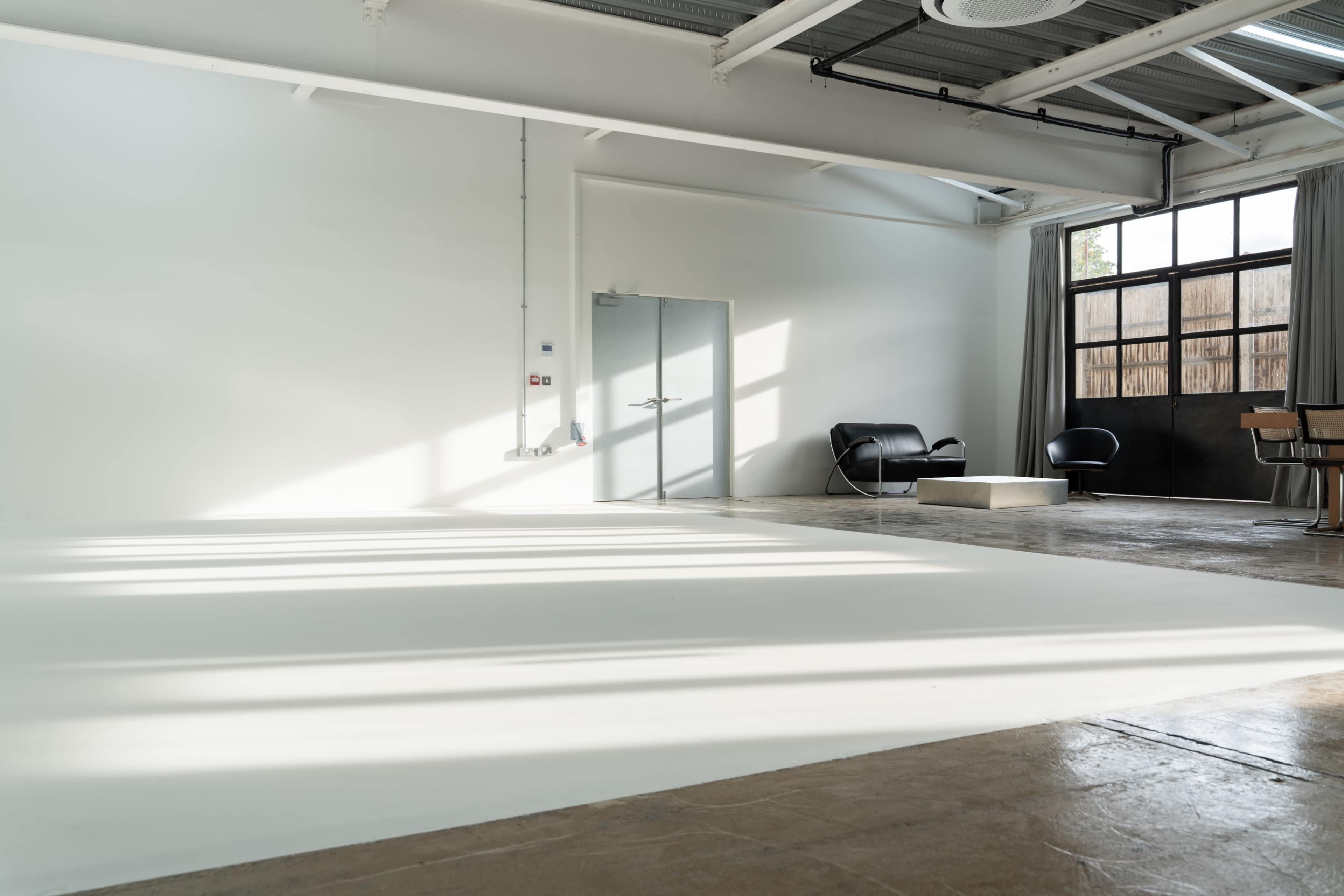 The image shows a spacious, well-lit room with a gray door, a large window, minimalist furniture, and a smooth white floor.