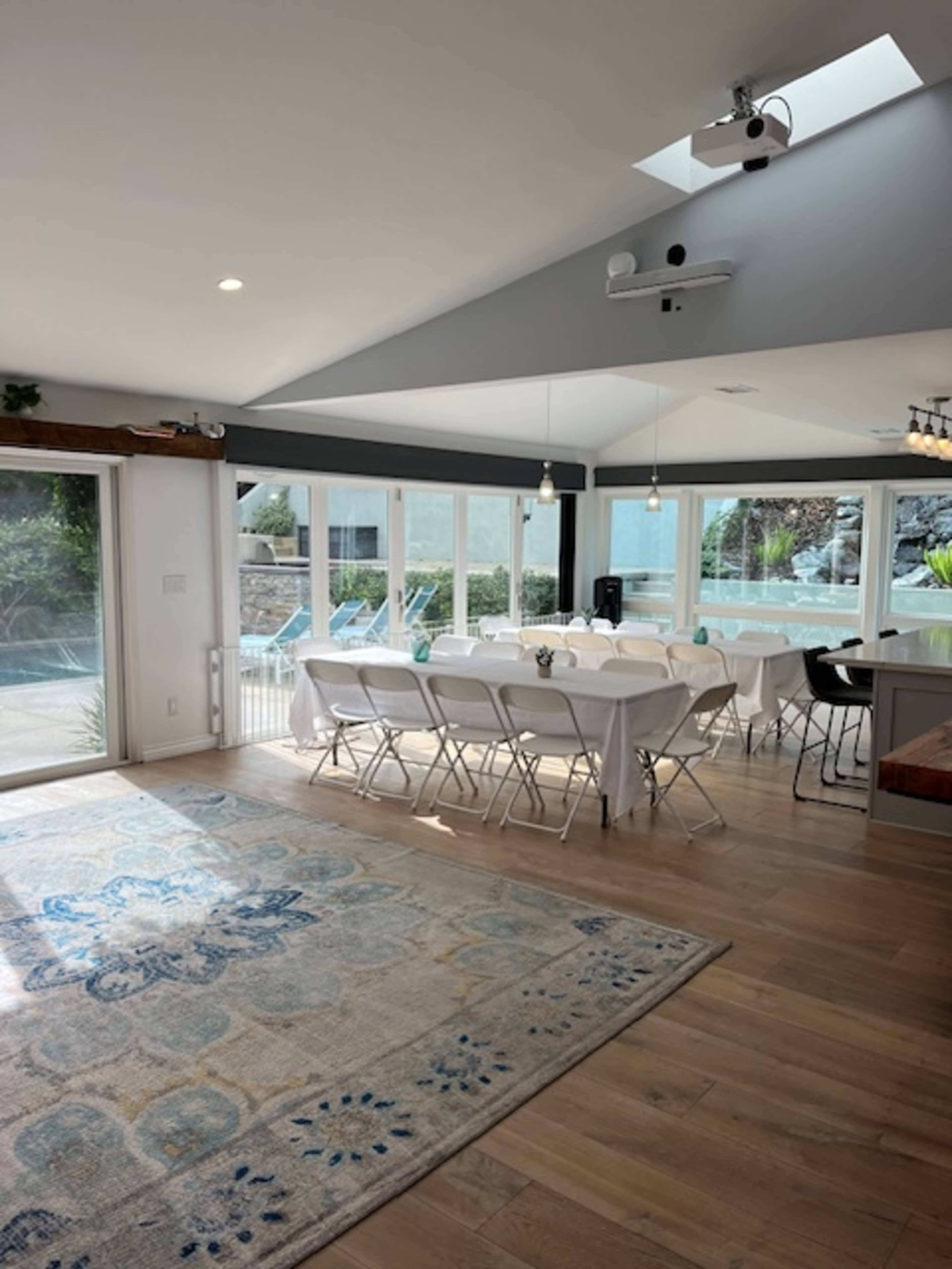 A spacious dining area features a long table set with chairs, large windows overlooking a pool, and a light-colored rug on the wooden floor.