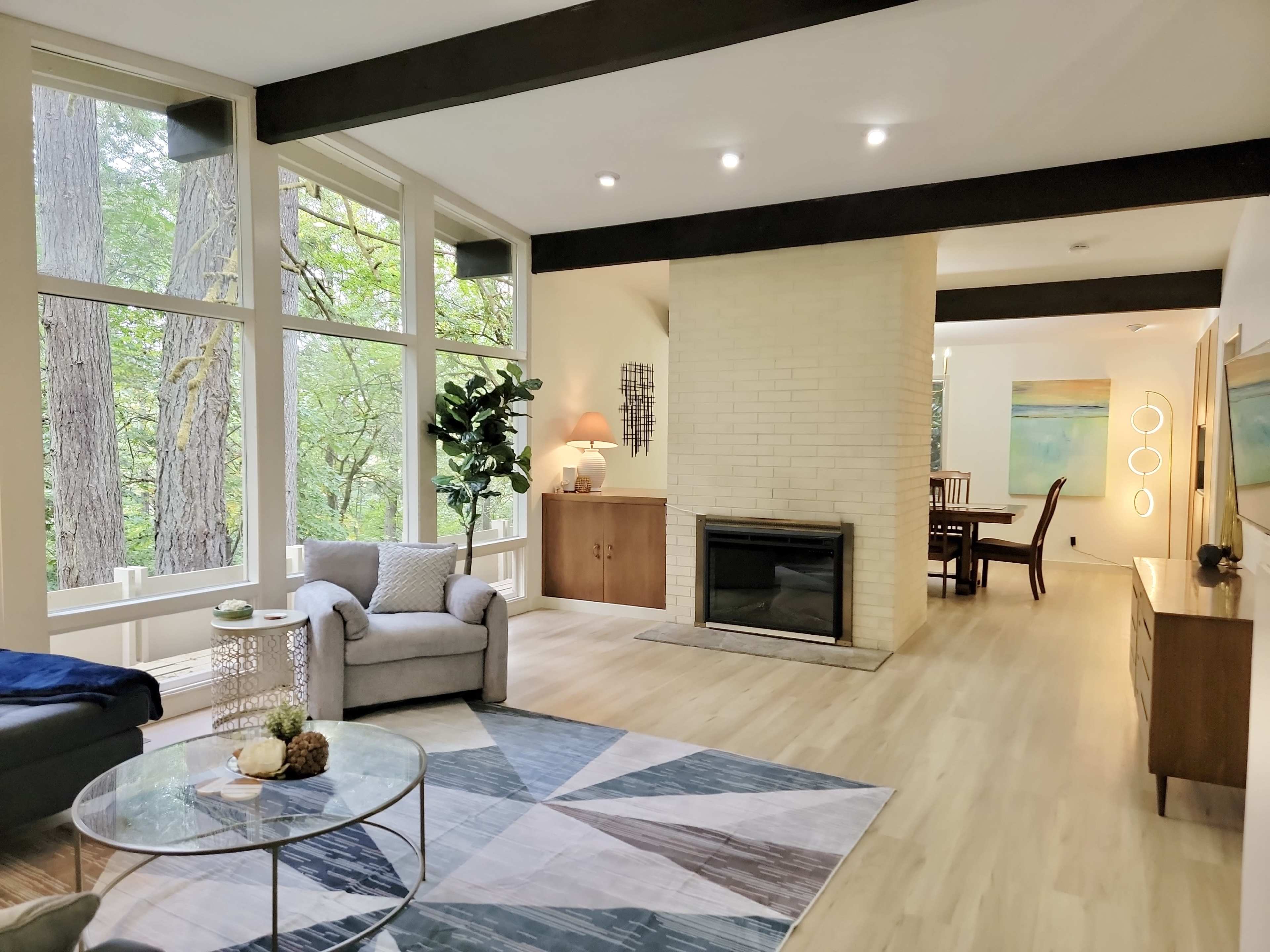 A modern living room features large windows with views of greenery, a fireplace, and a mix of contemporary furniture.