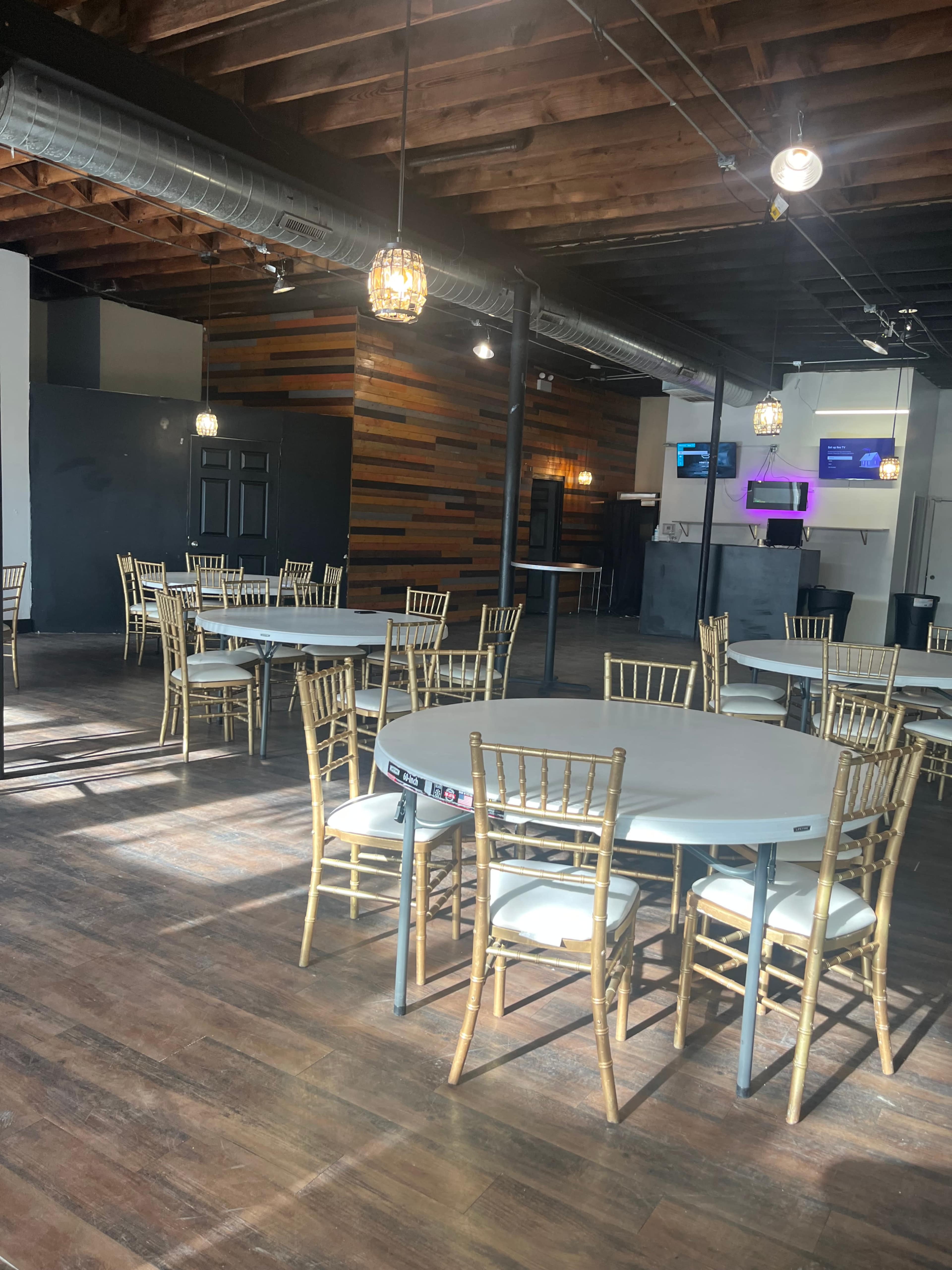 The image shows a spacious, empty event venue with wooden floors, several round tables with gold chiavari chairs, and a backdrop of wood paneling.