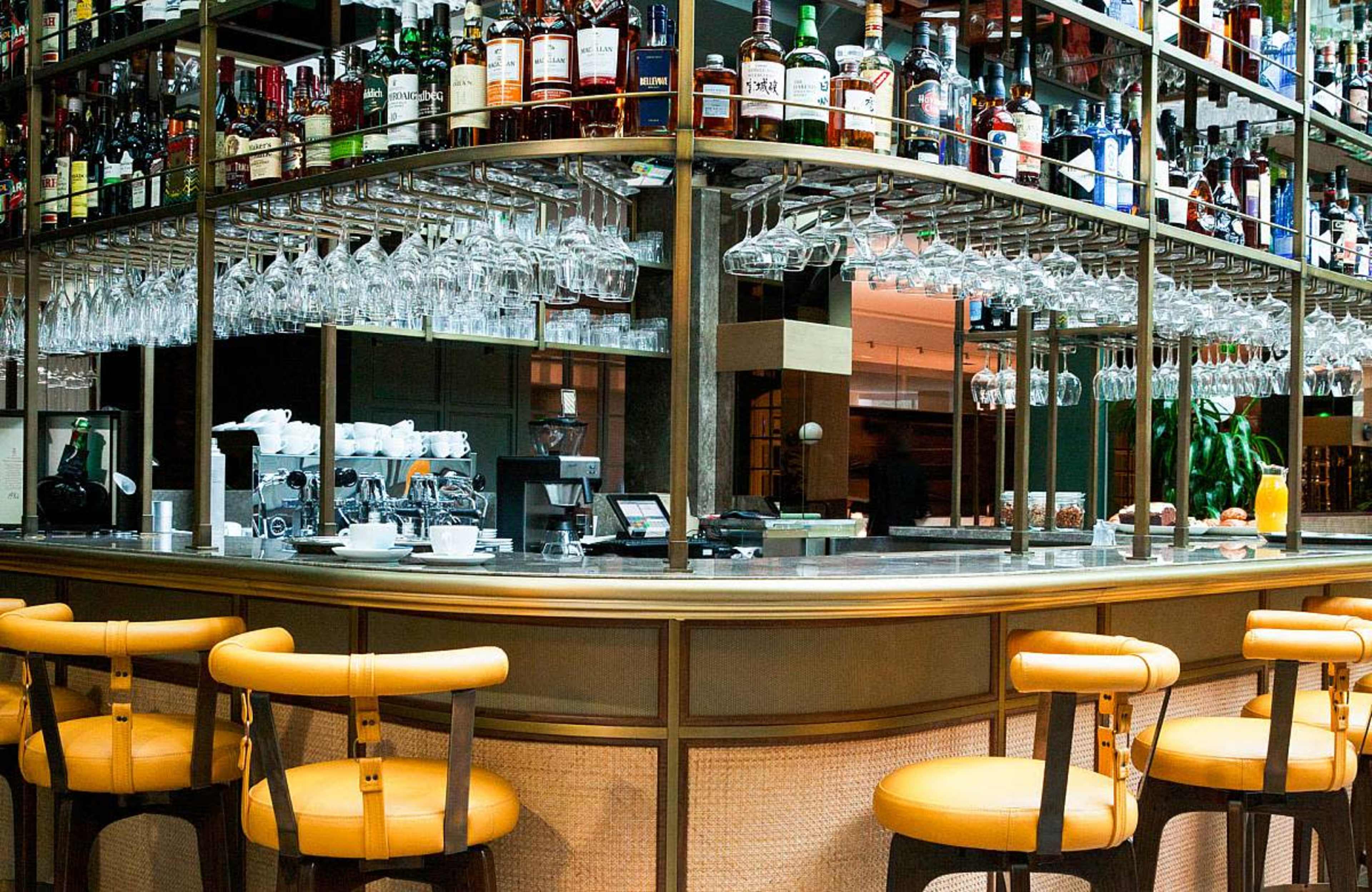 A bar area with a wide selection of liquor bottles on shelves above a counter, complemented by hanging wine glasses and several orange-cushioned chairs.