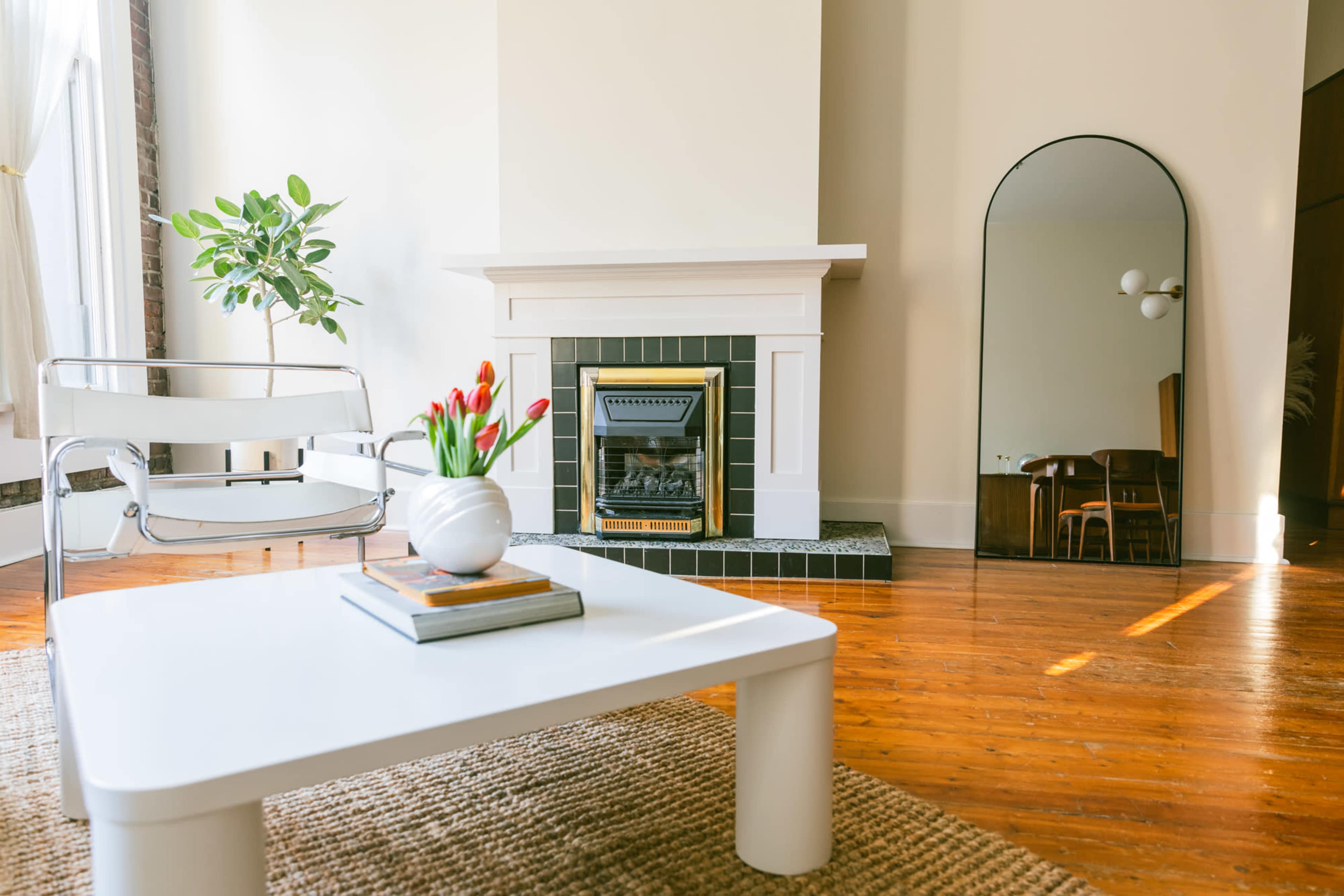A living room with a white coffee table, a plant near a fireplace, a large mirror, and a dining area in the background.